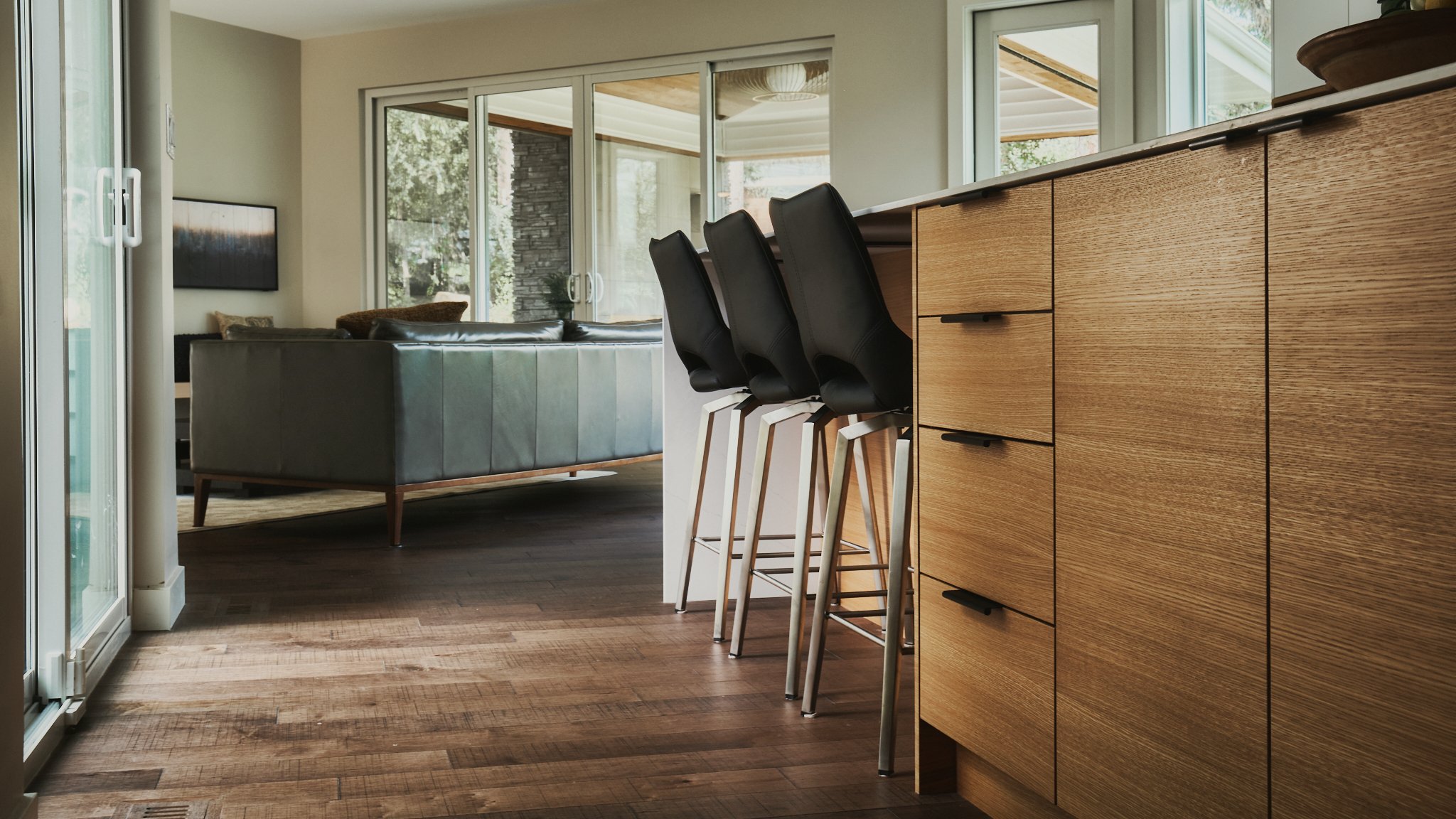 Modern open-concept living space featuring a kitchen with a wooden cabinet and three black barstools, a living room with a gray sofa, and large windows and sliding glass doors allowing natural light.