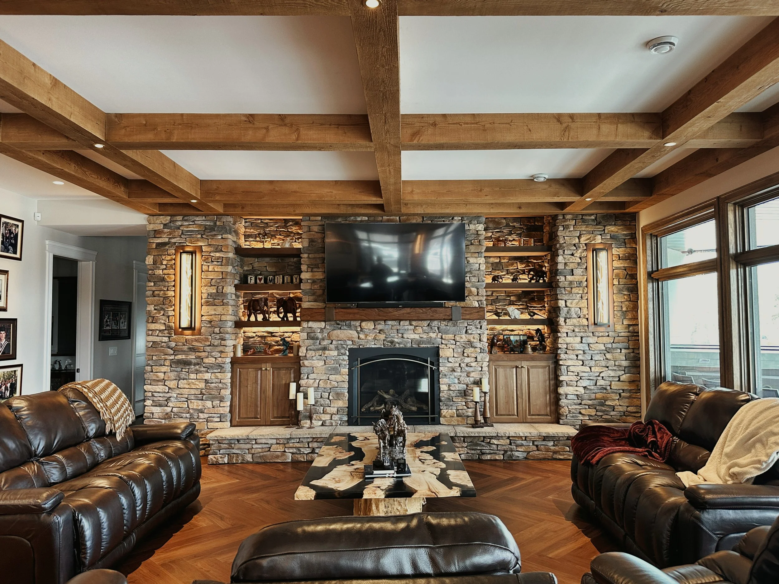 Living room with a stone fireplace, mounted flat-screen TV, wooden ceiling beams, large windows, leather sofas, and a black and white marble coffee table.