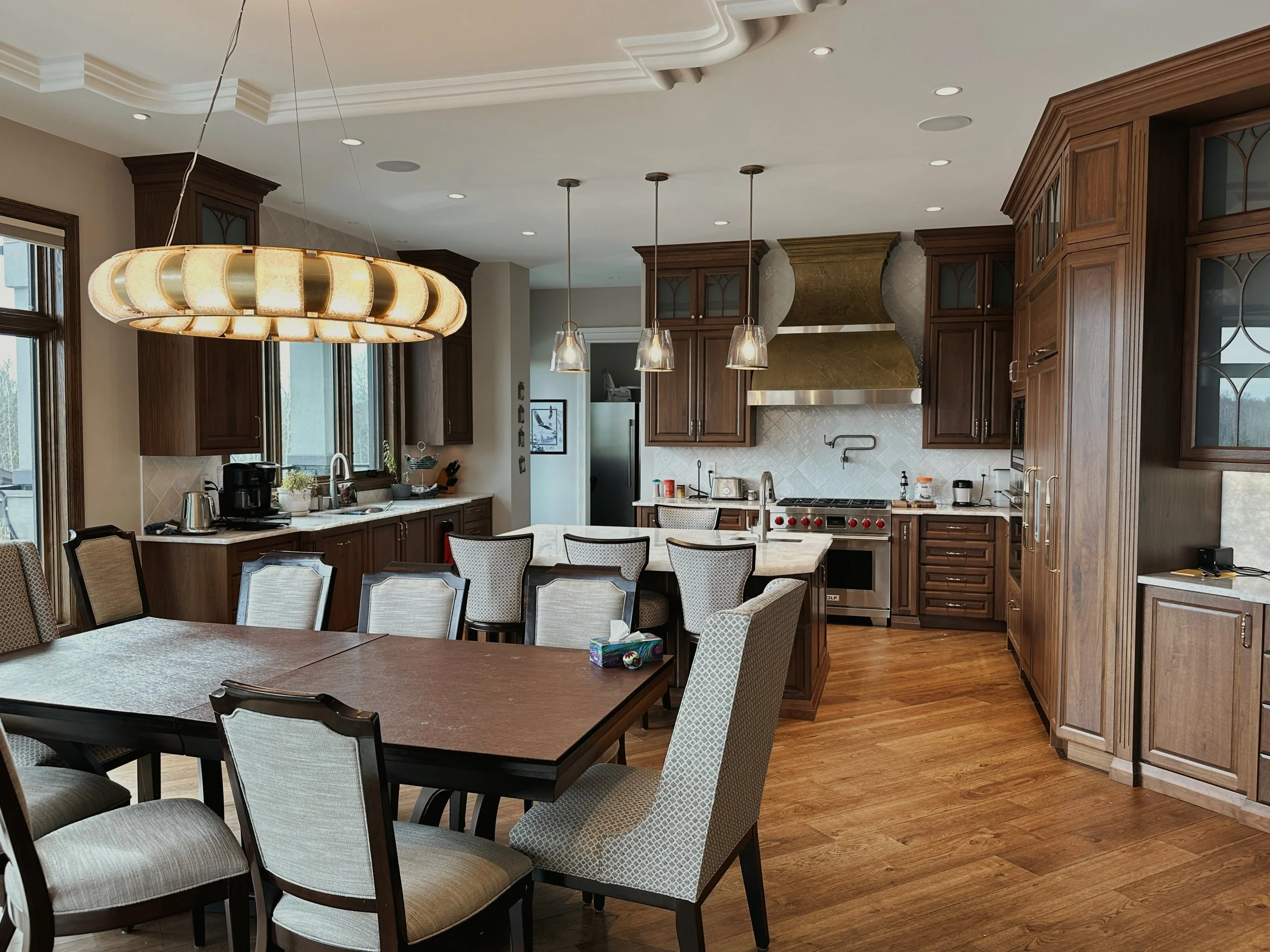 Open kitchen with wooden cabinets, stainless steel appliances, a large marble island, dining table with chairs, pendant and chandelier lighting, and hardwood floors.