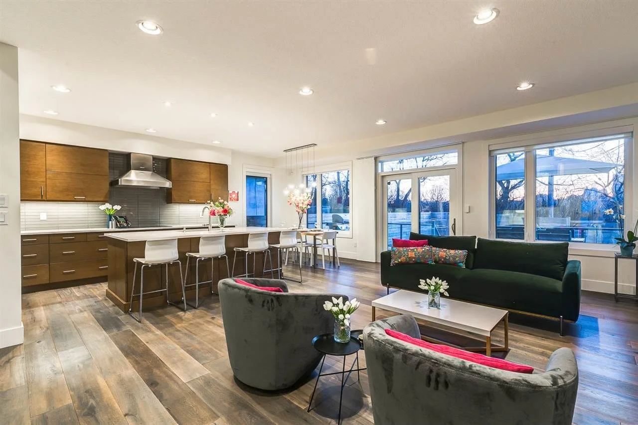 Open-concept living room and kitchen with hardwood floors, white walls, large windows, brown kitchen cabinets, white island with stools, black sofa with colorful cushions, and floral arrangements.