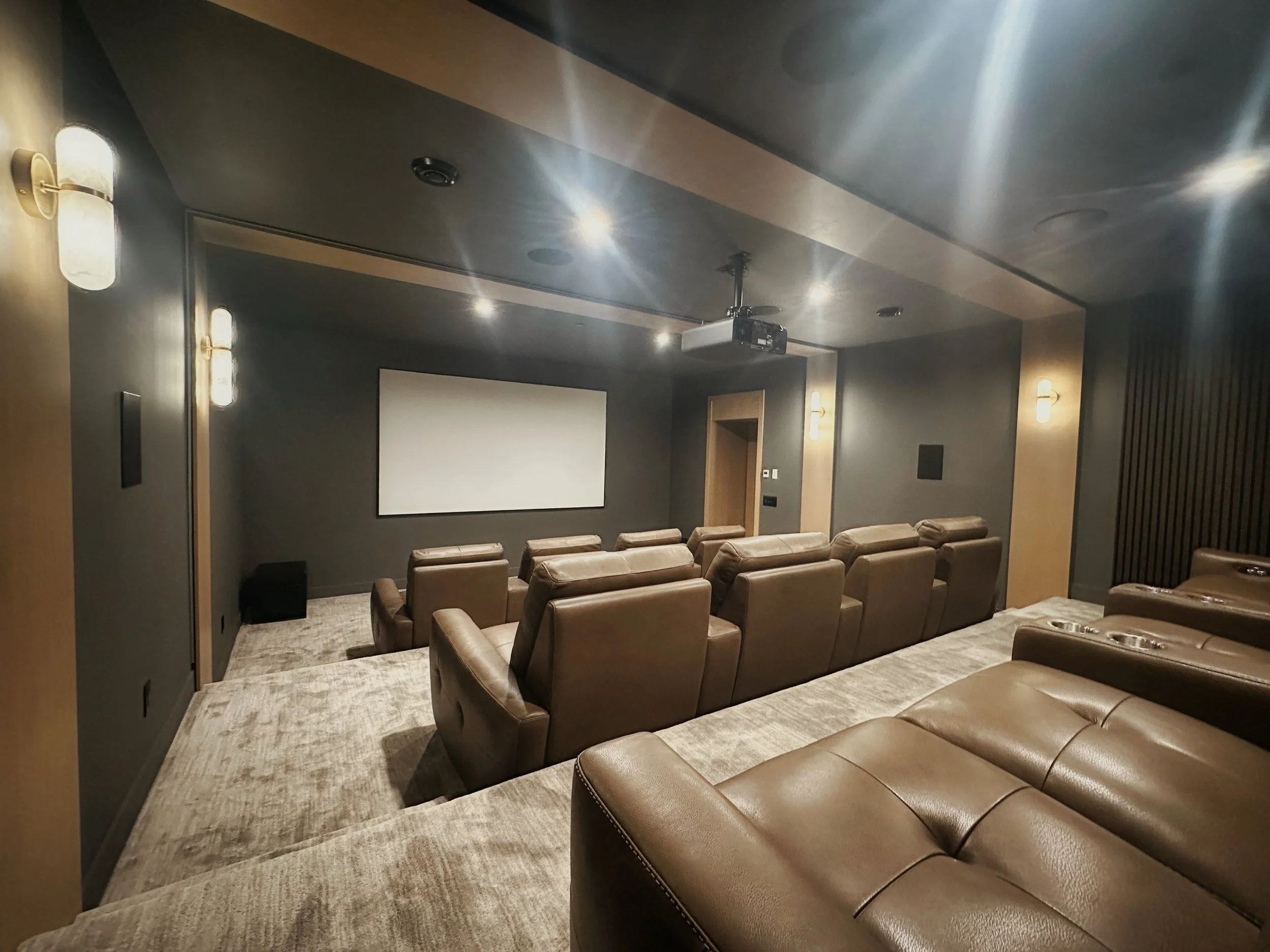 A modern home theater room with several brown recliner seats facing a large white projection screen, gray walls, carpeted floor, and wall-mounted lights.