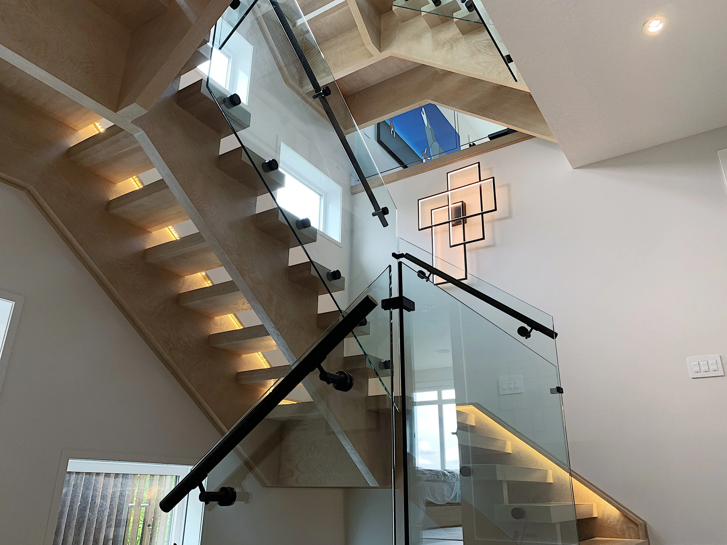 Modern staircase with light wood steps, integrated LED lighting, glass railings, and geometric wall fixture—custom design showcasing sleek architecture and ambient luxury interior style.