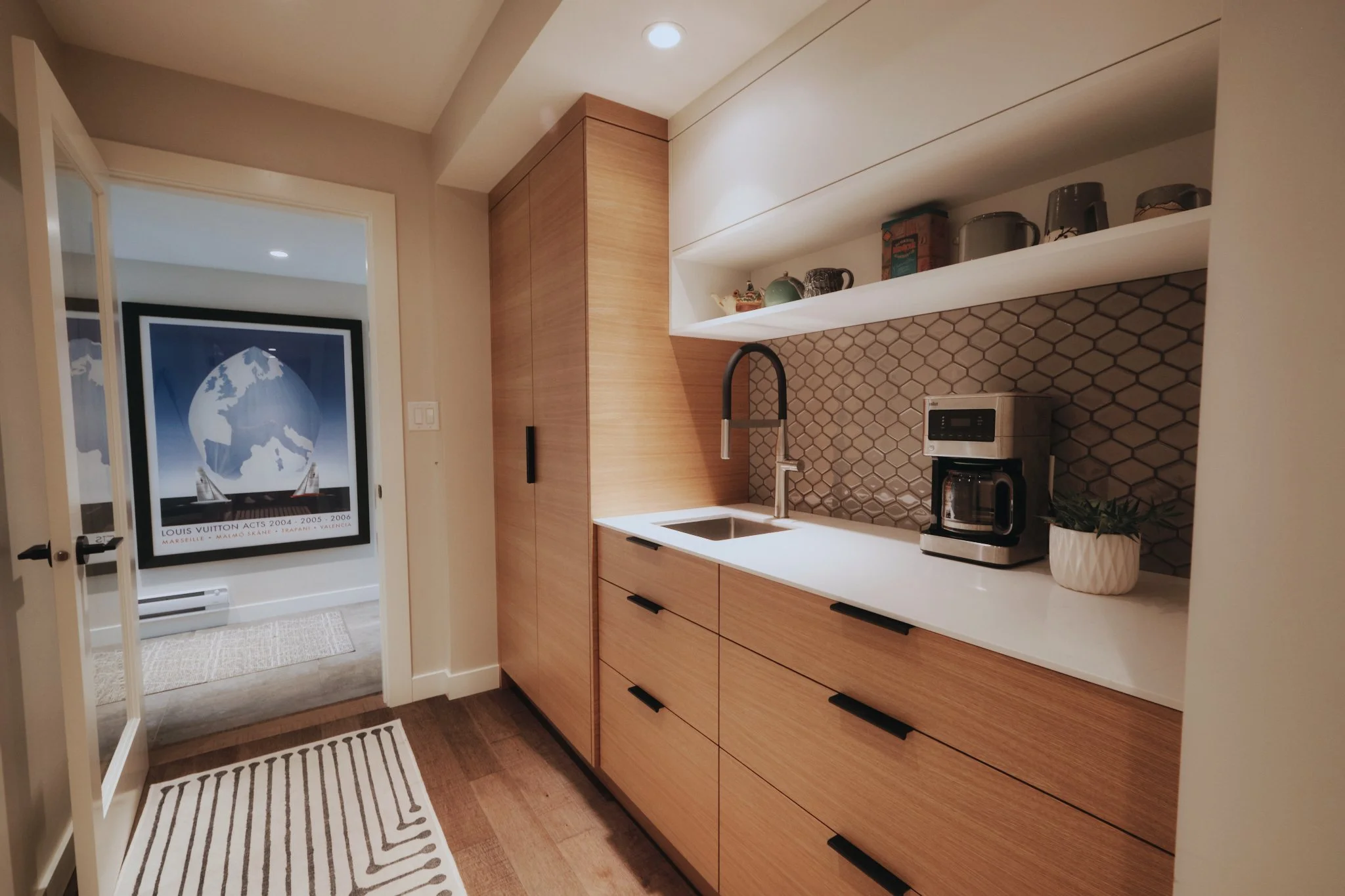 Modern kitchen with wood cabinetry, a white countertop, a coffee maker, a potted plant, and decorative items on open shelf. Hexagonal tile backsplash and a stainless steel sink with a black faucet. Bright lighting and a doorway leading to a room with