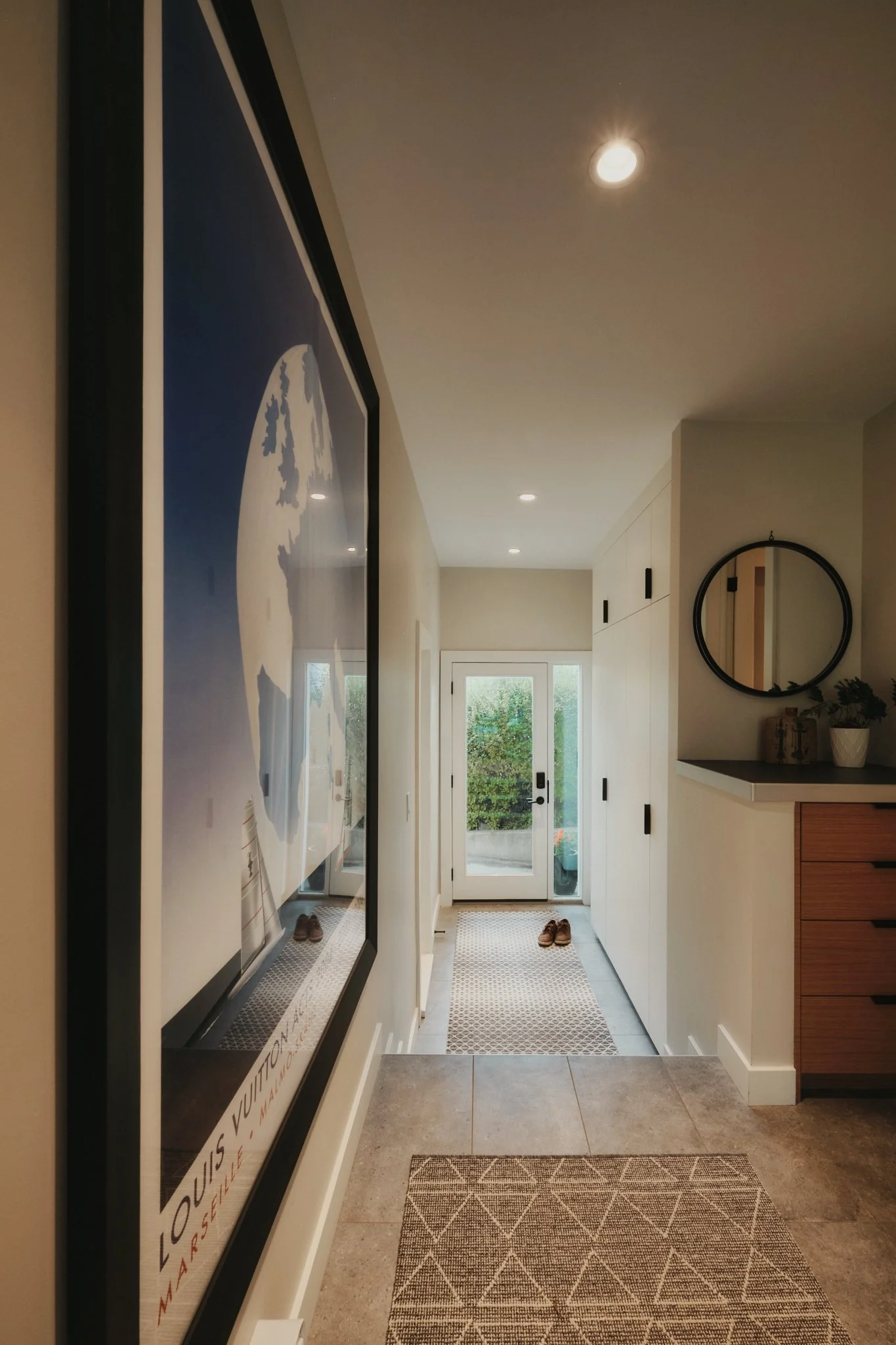 Entryway with glass door, two pairs of brown shoes, patterned rug, large framed art on the left wall, round mirror and potted plant on the right side near the wall.
