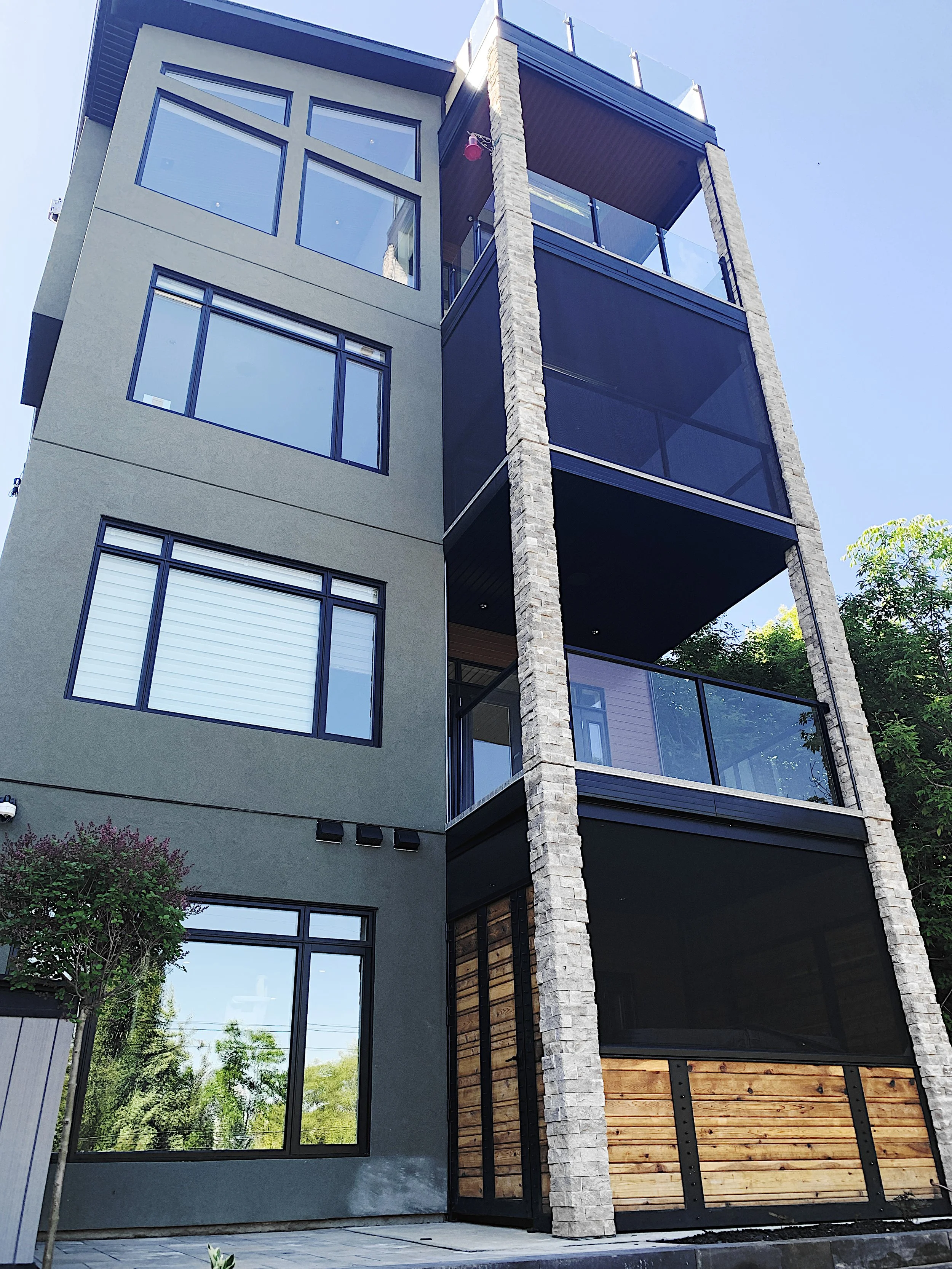 Multi-story residential home with large black-framed windows, stacked glass balconies, stone columns, and muted green-gray exterior—custom home builder design blending sleek and rustic elements.