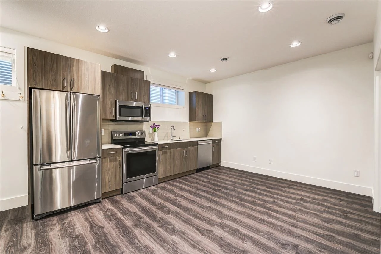 Empty kitchen in a secondary suite with stainless steel refrigerator, microwave, and stove, dark wood cabinets, a small window, and vinyl plank flooring.