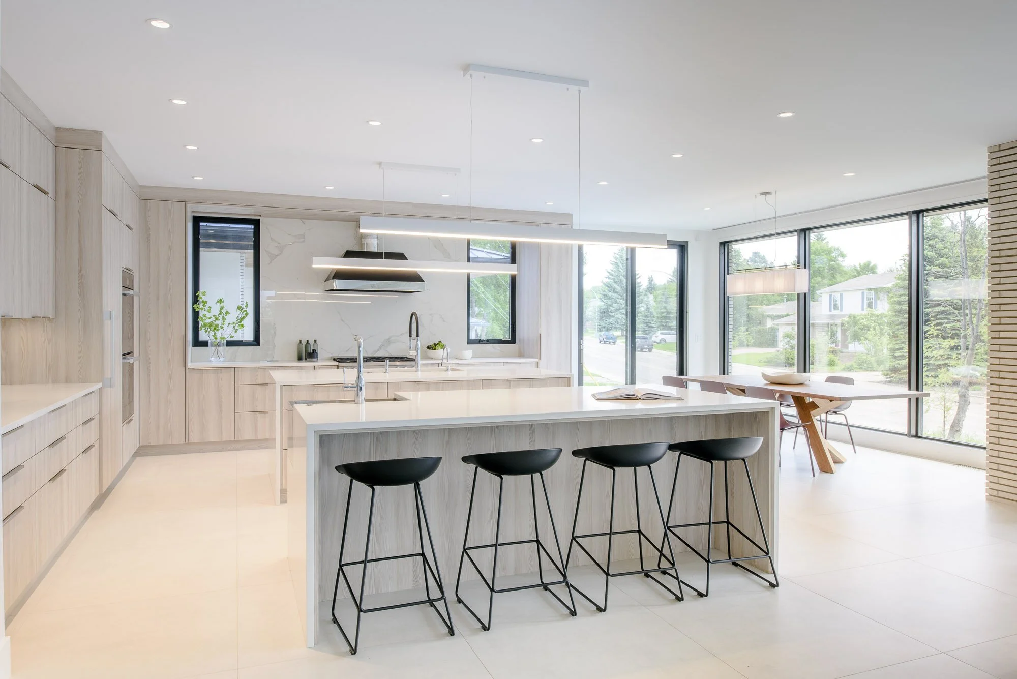 Modern open-concept kitchen with large white marble island, black counter stools, linear pendant lights, and floor-to-ceiling windows overlooking backyard.