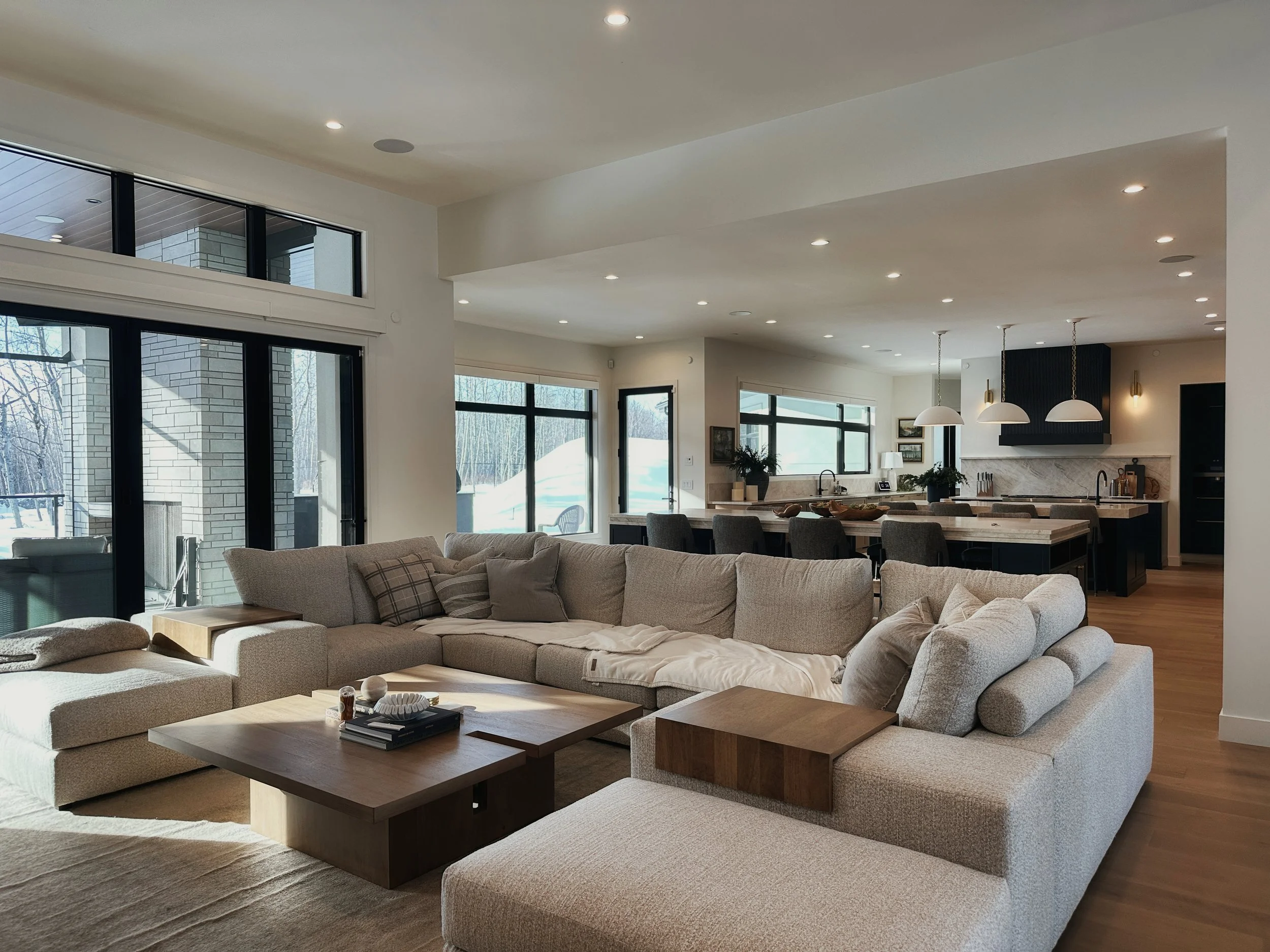 Spacious open-concept living room and kitchen with large sectional beige sofa, wooden coffee table, vaulted ceilings, floor-to-ceiling black-framed windows overlooking snowy forest, modern pendant lights, and connected marble island kitchen
