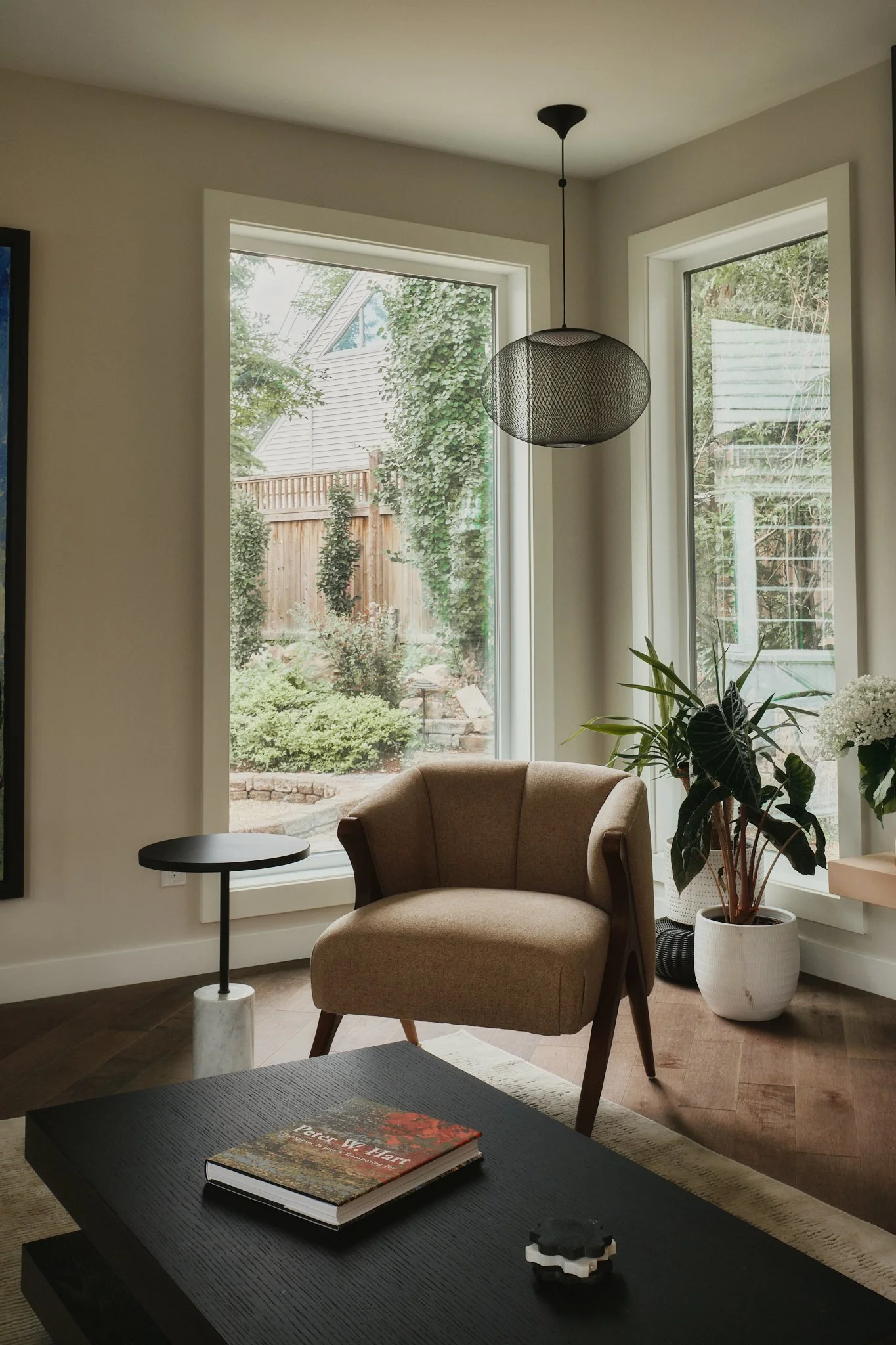 Cozy living room corner with mid-century armchair, pendant light, coffee table, and garden view through large windows.