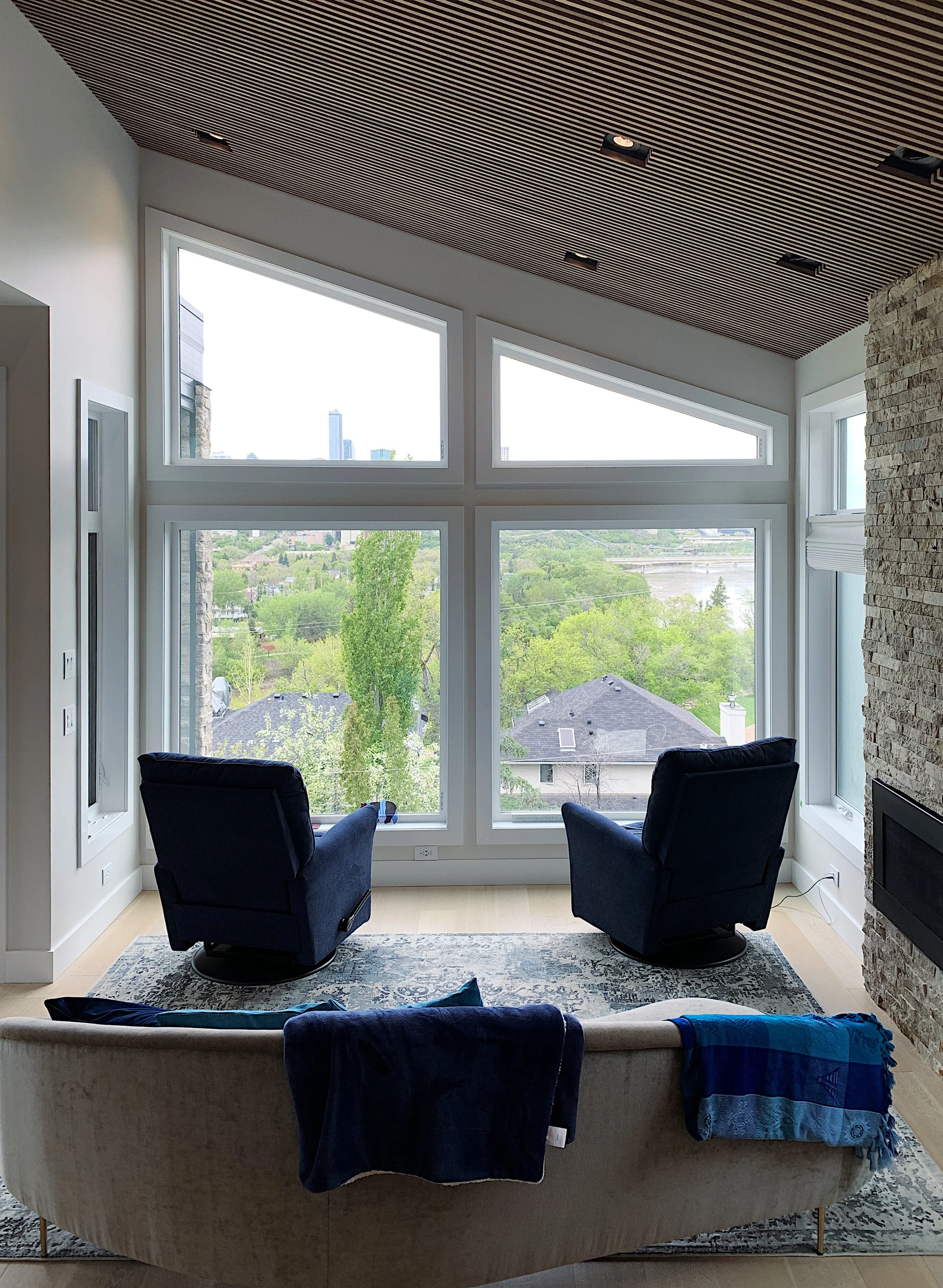 Living room with large rake window showcasing lush landscape and river view—features dark blue armchairs, curved beige sofa, wood slat ceiling, stone fireplace, and serene natural lighting.