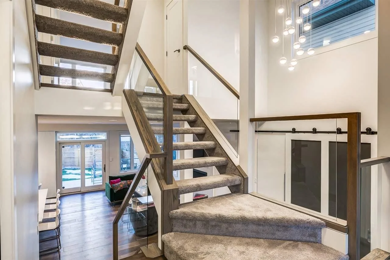 Interior view of a modern home staircase with carpeted steps and wooden railings, leading to an upper floor, with a glimpse into a living room with glass doors and wooden flooring.