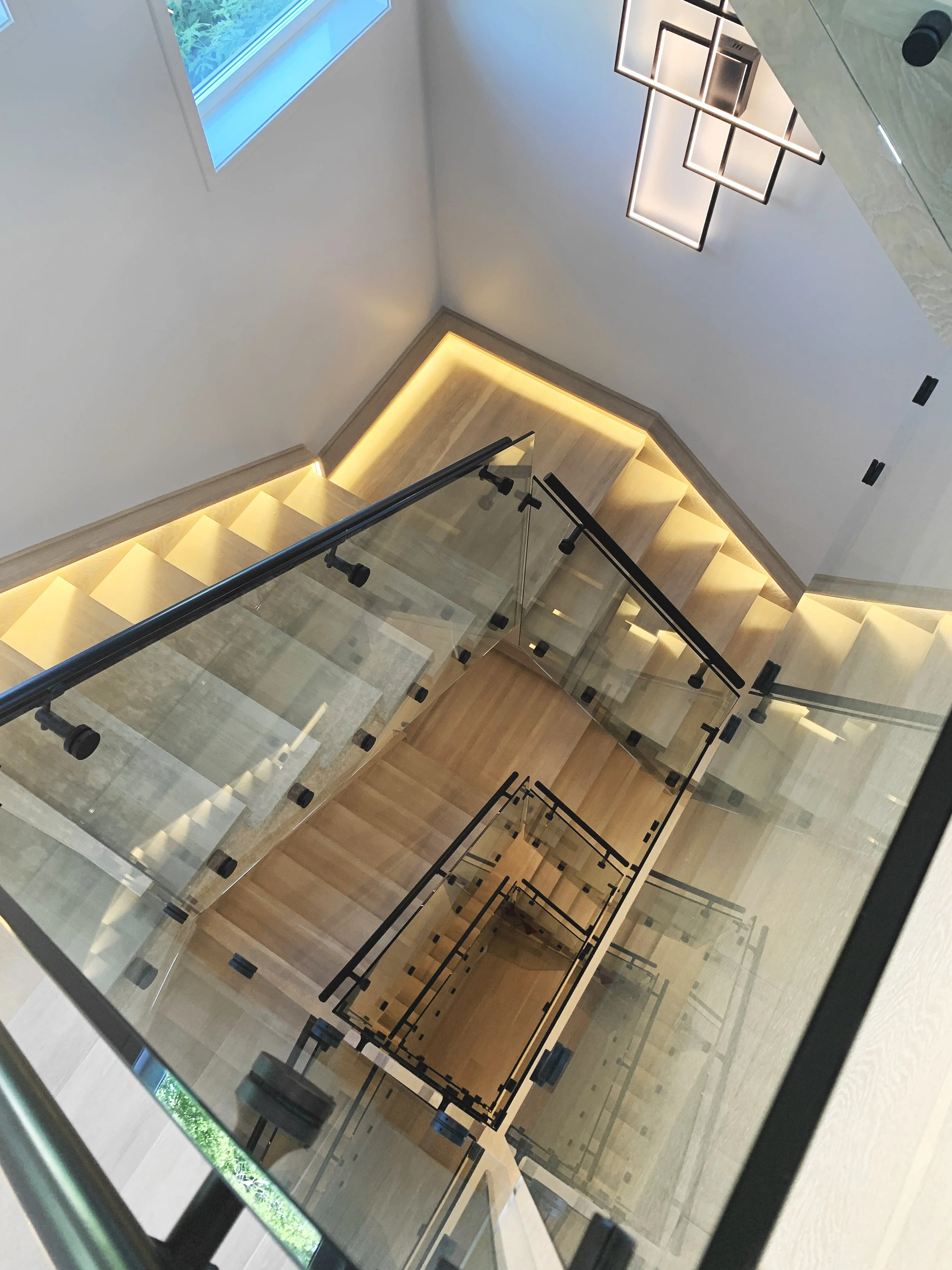 Modern staircase with light wood steps, glass railings, and black metal supports—features geometric spiral design, ambient lighting, and contemporary chandelier ideal for custom home builder luxury interiors.