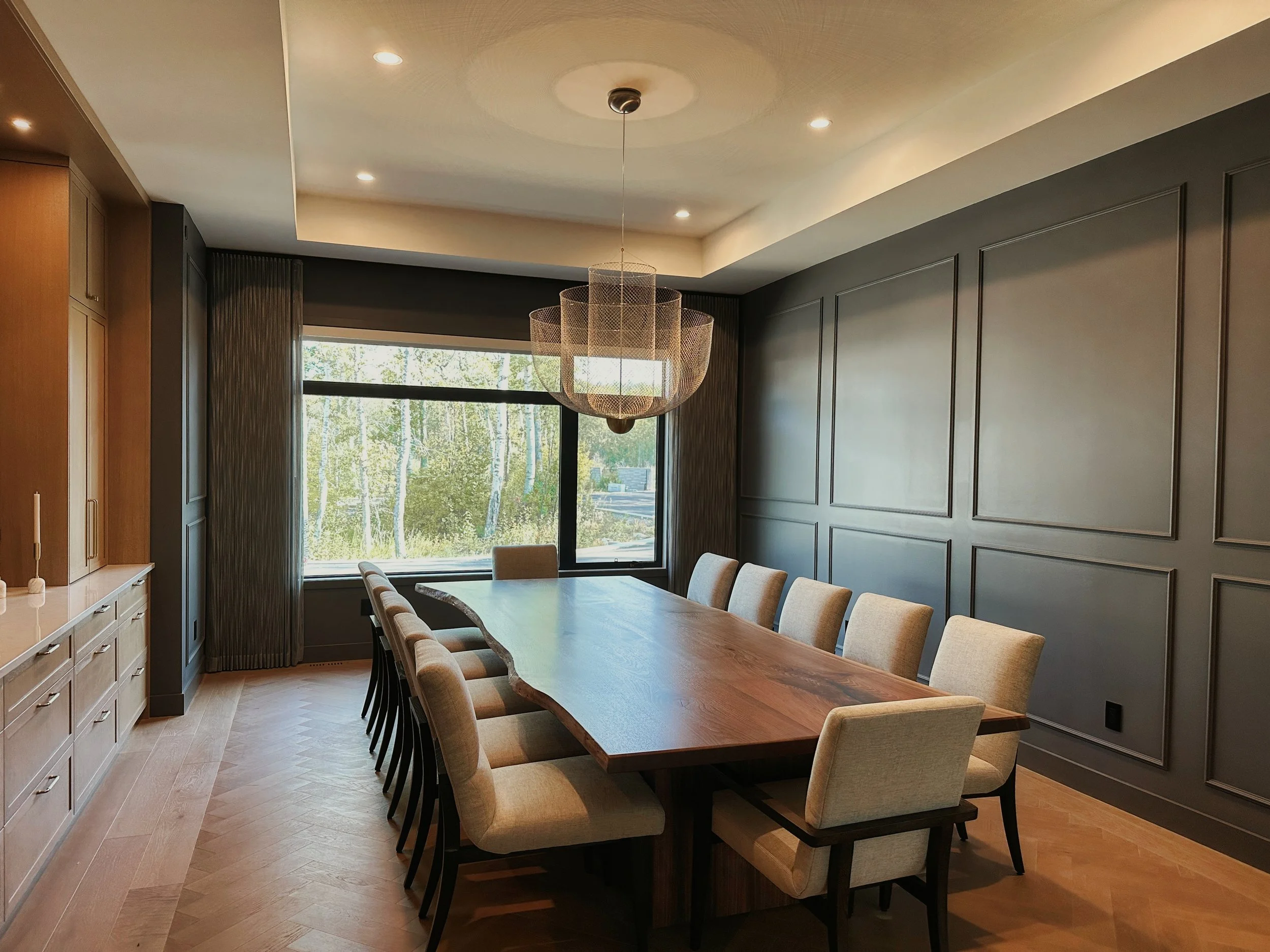 Sophisticated modern dining area with natural-edge walnut table seating ten, light beige upholstered chairs, woven orb pendant, expansive forest window, grey geometric panels, herringbone hardwood.