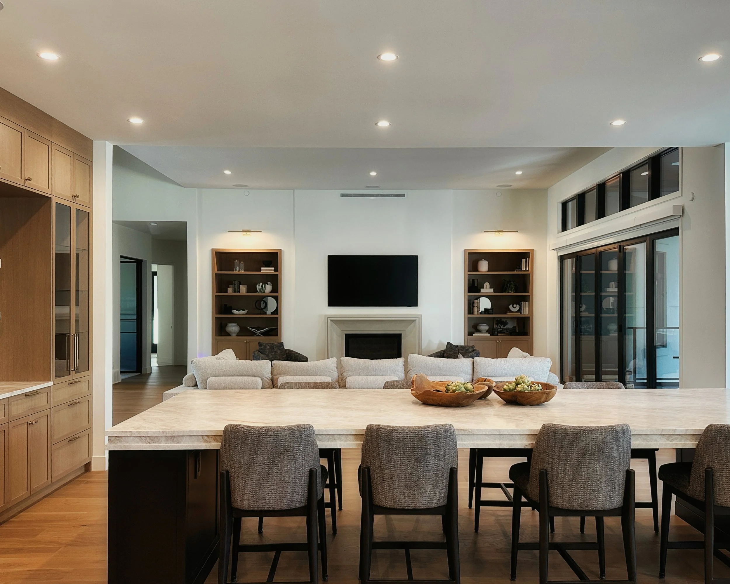 Modern open-concept great room with large marble island, grey bar stools, walnut cabinetry, large sectional sofa, stone fireplace, built-in bookshelves, recessed lighting, and neutral luxury interior.