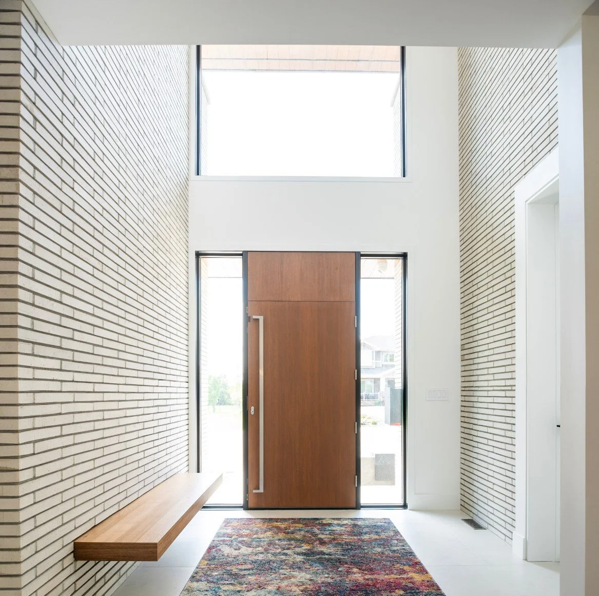 Sleek contemporary foyer design with vaulted ceiling, oversized wooden front door and glass panels, neutral brick texture walls, integrated wooden bench seat, bright overhead daylight, and artistic vibrant floor rug.