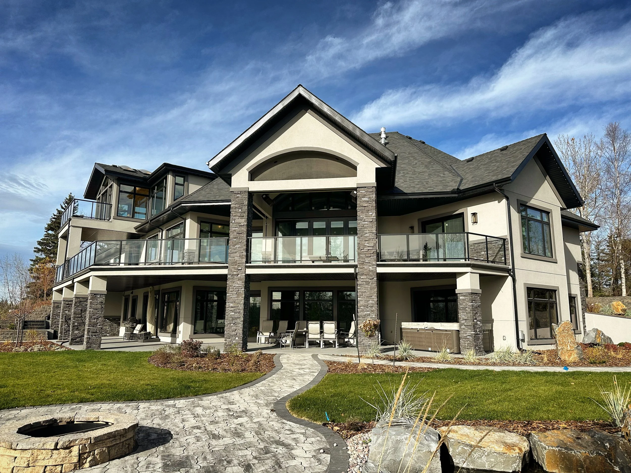 Large modern house with multiple balconies, glass railings, and stone accents, surrounded by a landscaped yard with a paved stone pathway and rocks.
