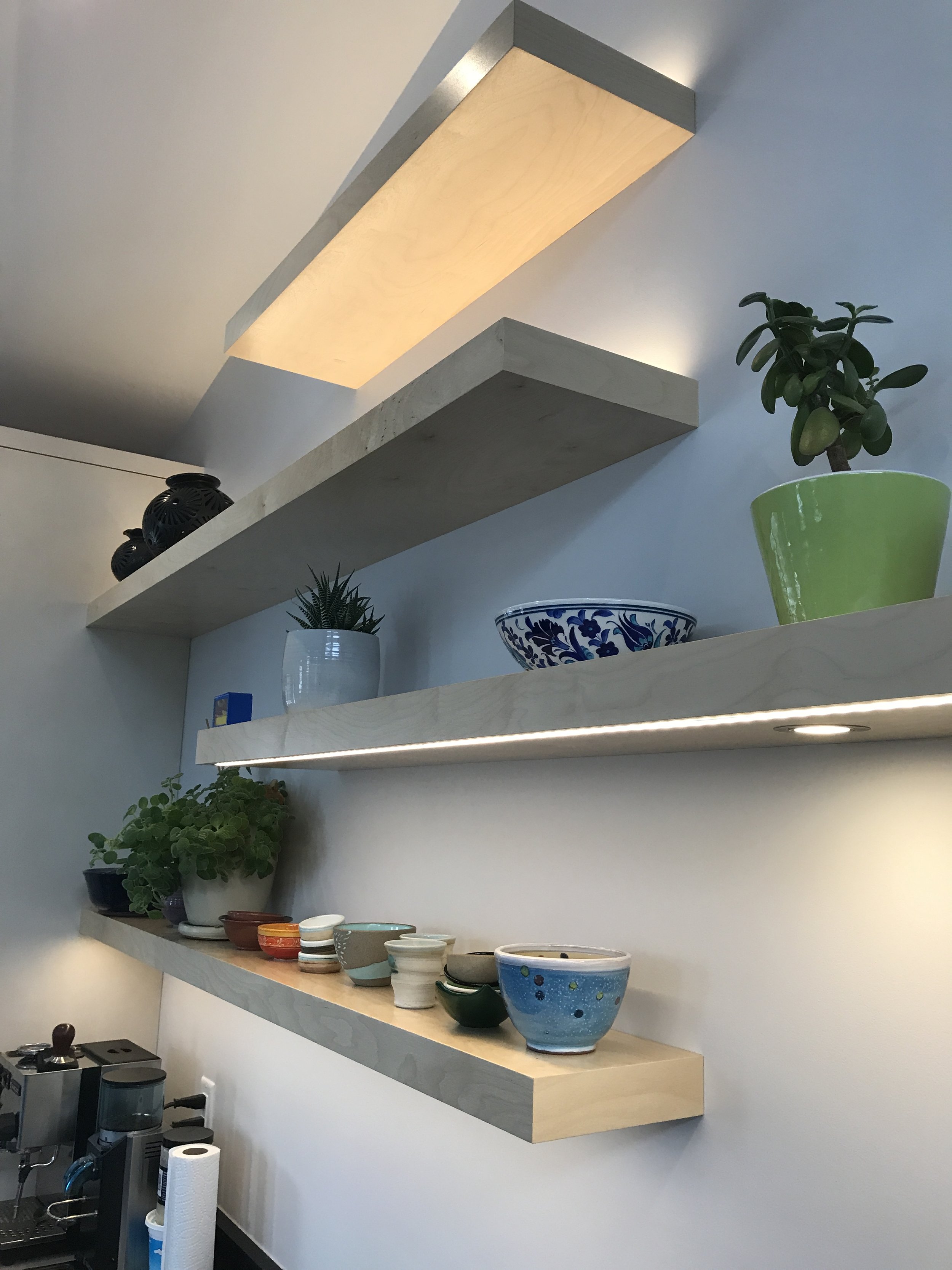 Modern kitchen wall with three wooden floating shelves featuring integrated lighting, ceramic dishes, potted plants, and a coffee machine below—minimalist design ideal for stylish home interiors.