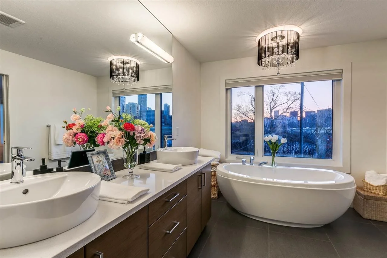 Modern bathroom with double vanity sinks, a free-standing bathtub, and city skyline view through the windows. Decor includes flowers and framed photos.