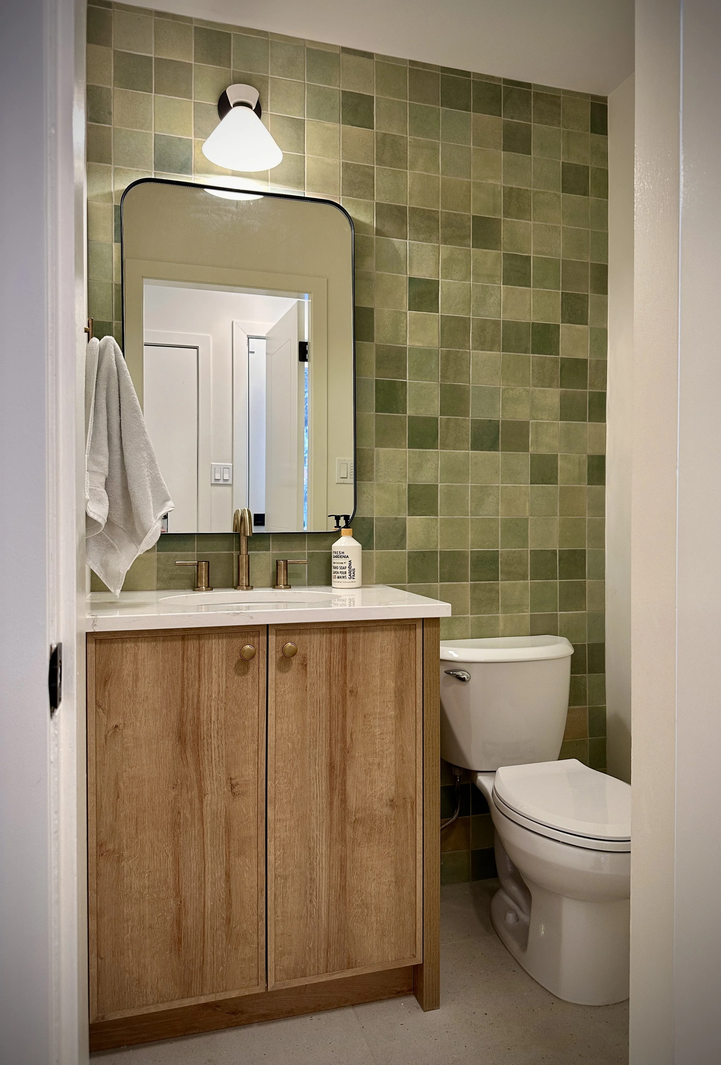 A small bathroom with a green tiled wall, a wooden vanity with a white countertop, a rectangular mirror, a wall-mounted light fixture, a soap dispenser, a white towel hanging on a hook, and a white toilet.