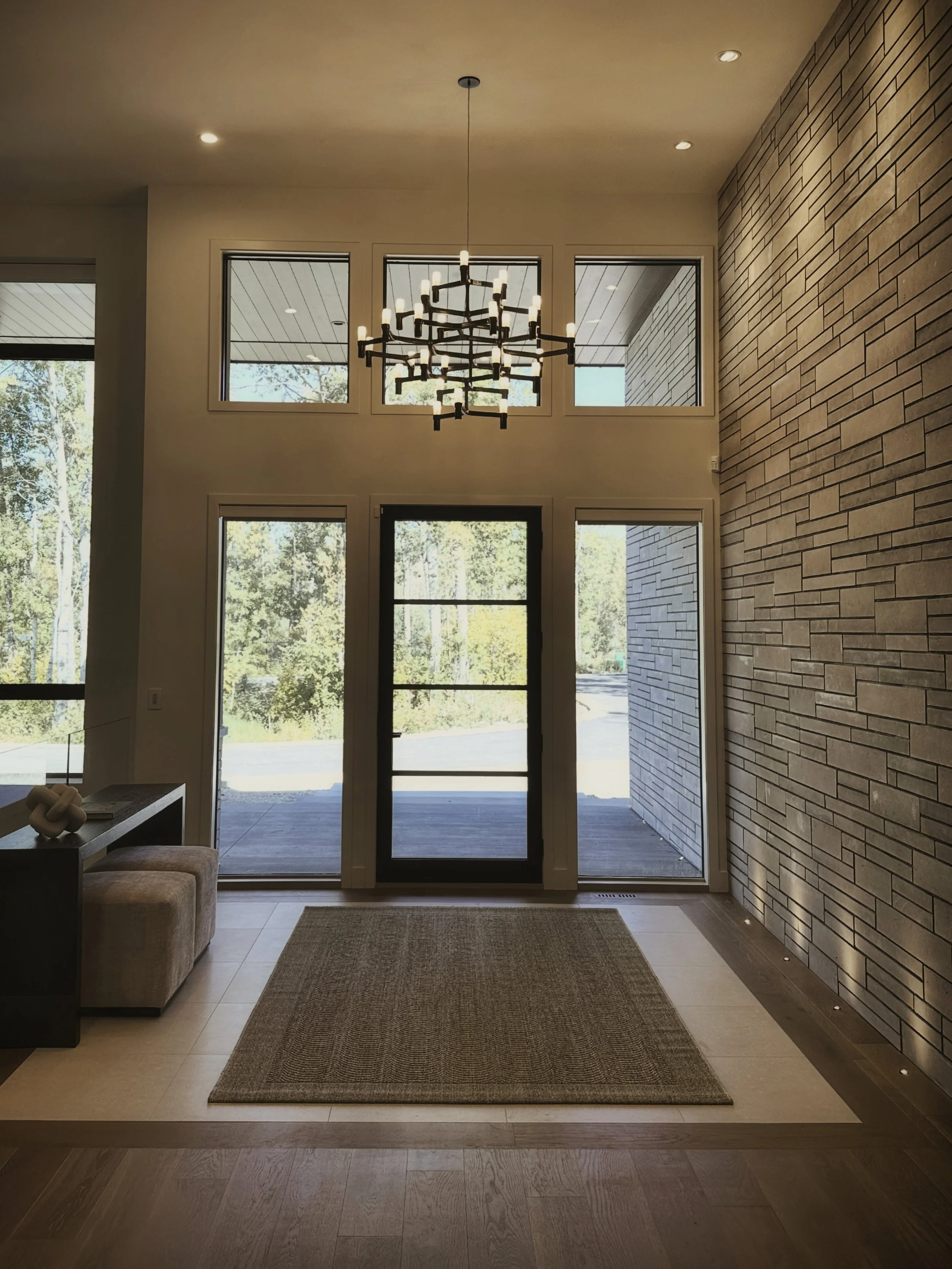High-end two-story entryway with geometric black chandelier, textured grey stone cladding, expansive black front door and sidelights, triple transom windows above displaying aspen grove, neutral walls, recessed lighting, woven neutral rug.