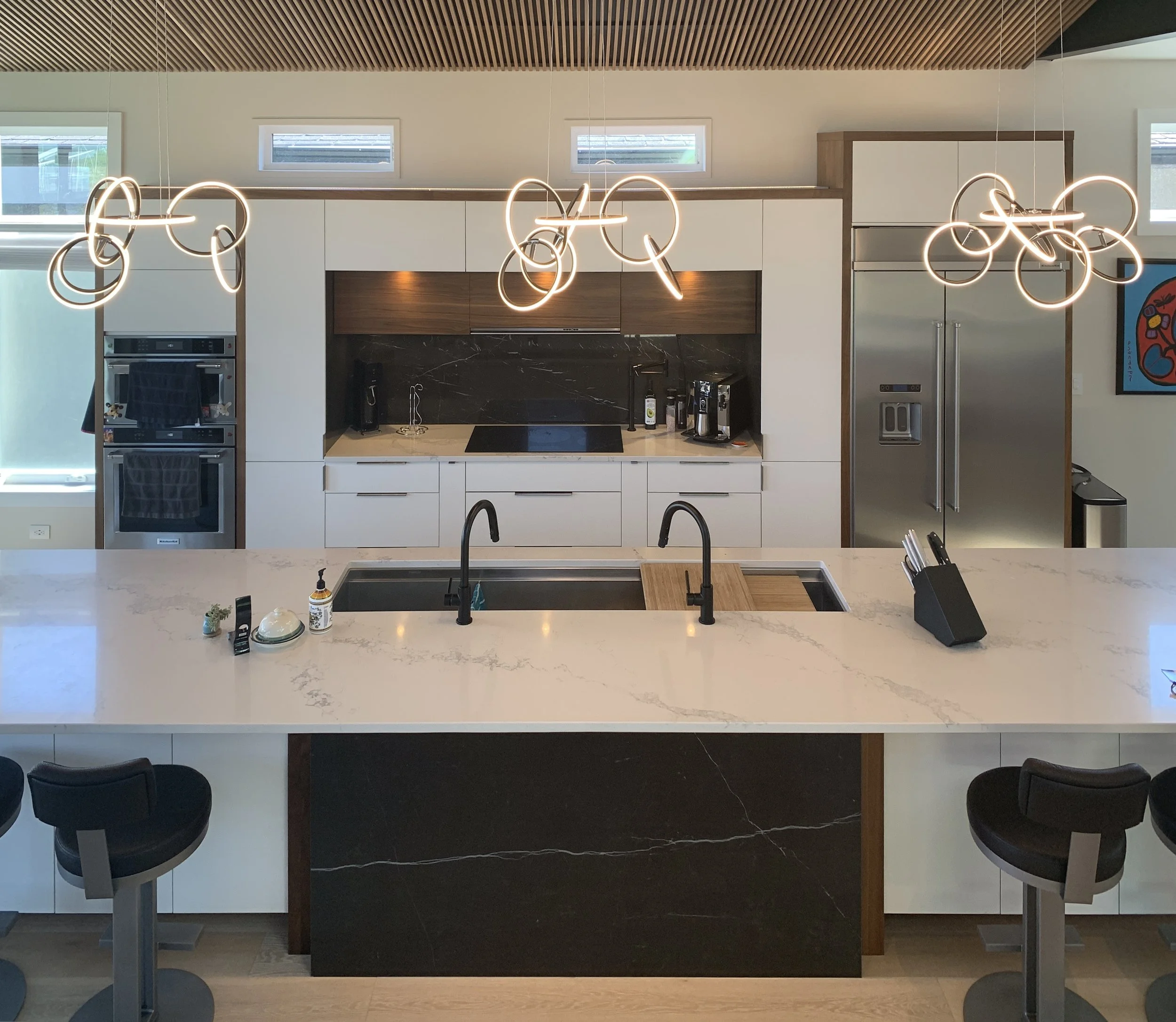 Modern kitchen with white marble island, black base, double sink, looped pendant lights, wood-slat ceiling, and built-in cabinetry