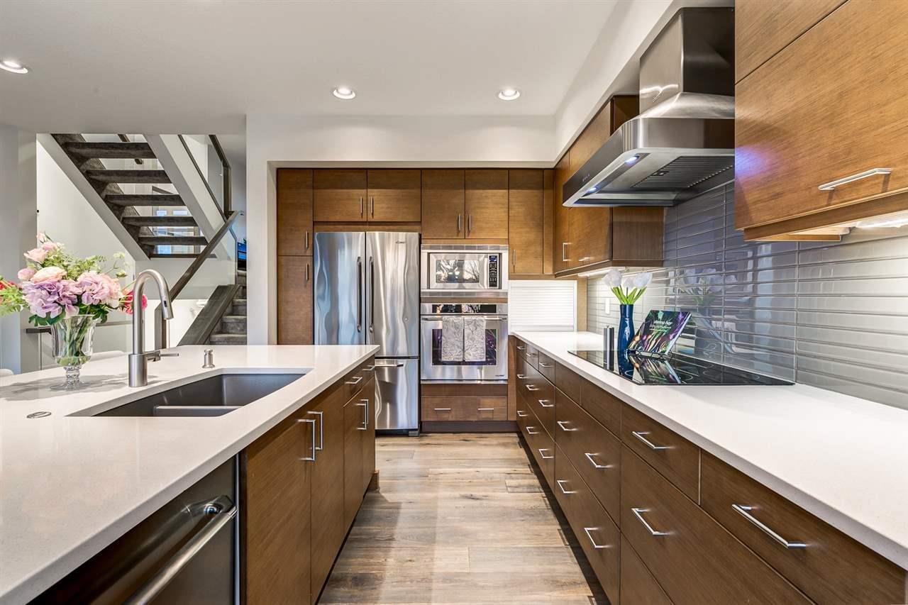 Modern kitchen with wooden cabinets, white countertops, stainless steel appliances including a refrigerator and microwave, a cooktop, a sink with a flower arrangement, and a staircase seen in the background.