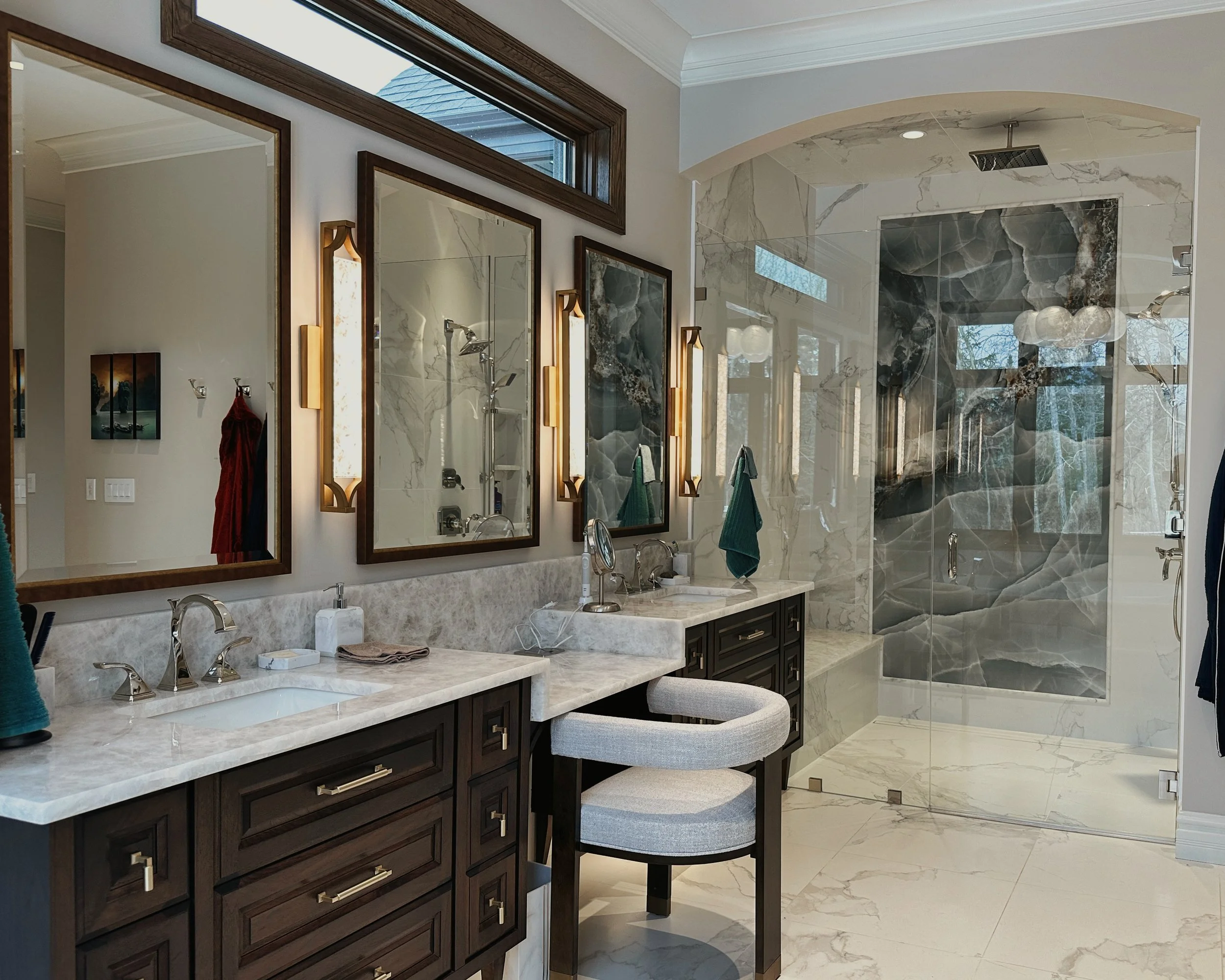 Luxury bathroom with dual vanities, large mirrors, marble countertops, modern light fixtures, and a glass-enclosed walk-in shower with marble walls and a rainfall showerhead.
