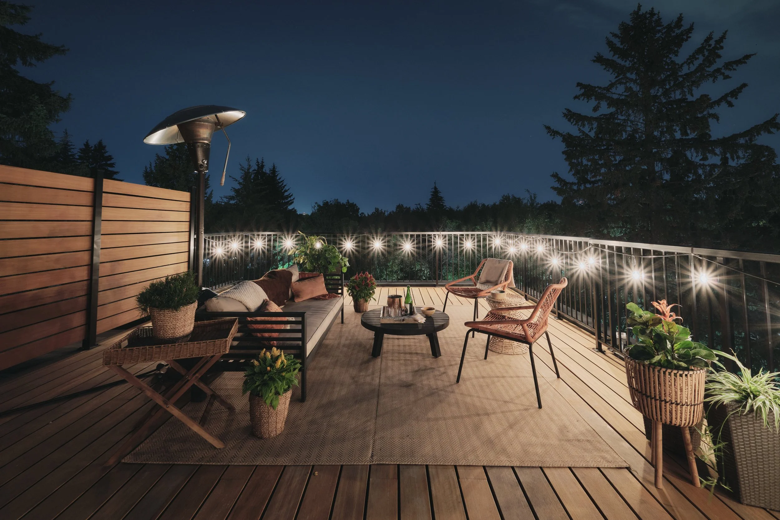 Contemporary deck featuring wood flooring, black railing, and illuminated privacy wall under night sky.