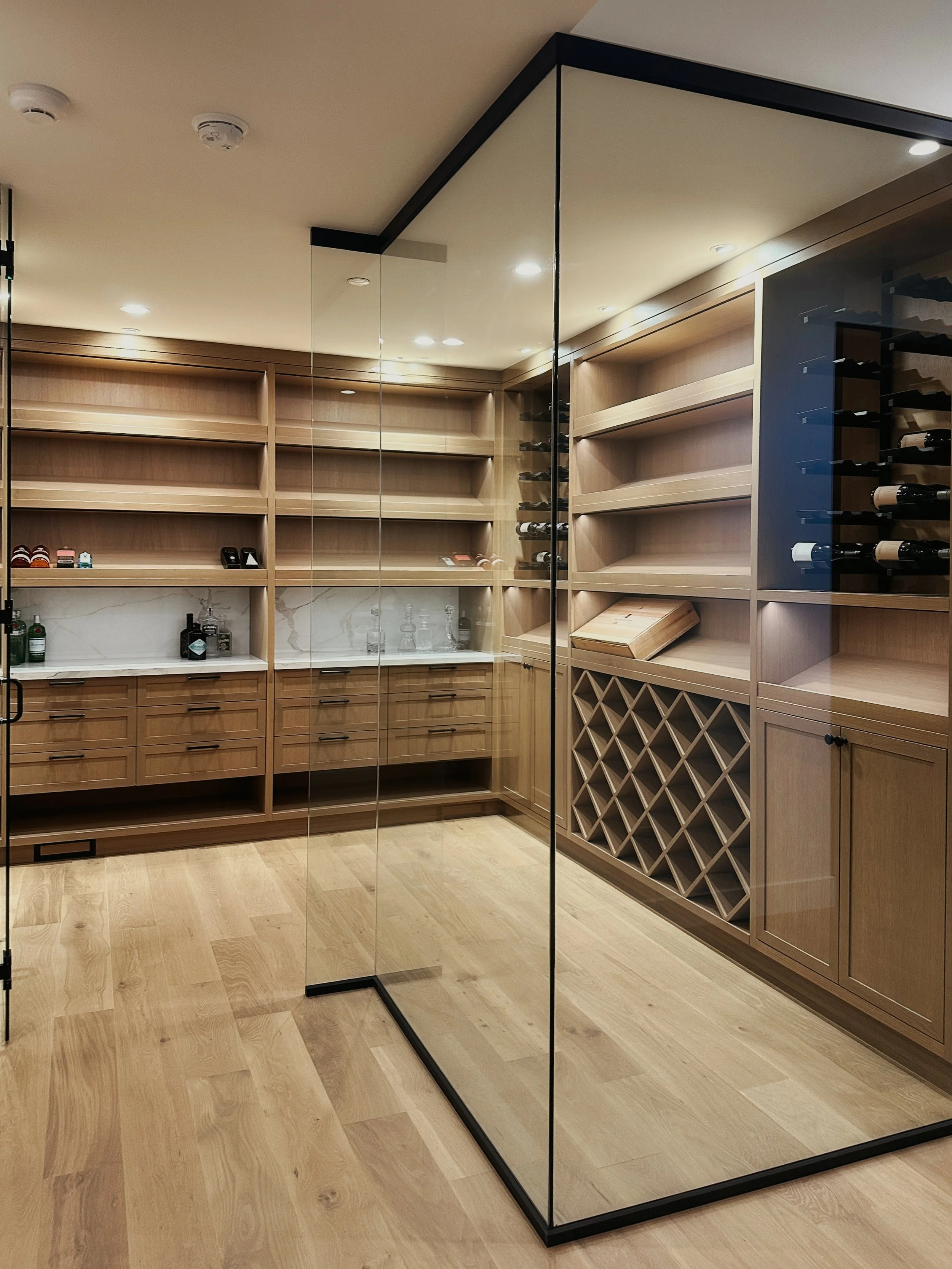 Modern wine cellar with wooden shelves, wine racks, cabinets, and a glass partition, featuring bottles, drawers, and bar accessories on a marble countertop.