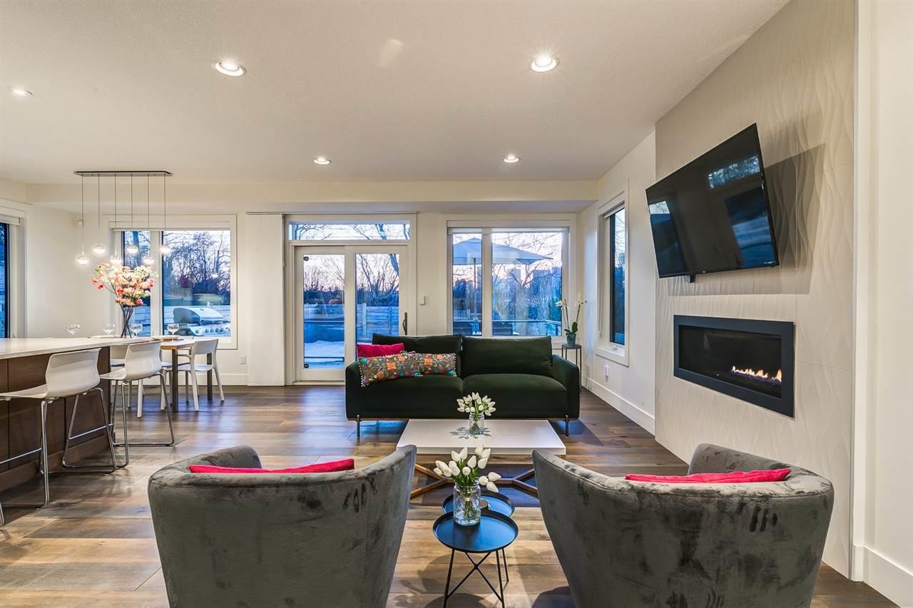 Modern living room with a green sofa, two gray velvet armchairs, a black coffee table with flowers, a white floating fireplace, a large flat-screen TV, and floor-to-ceiling windows that show a winter landscape outside.