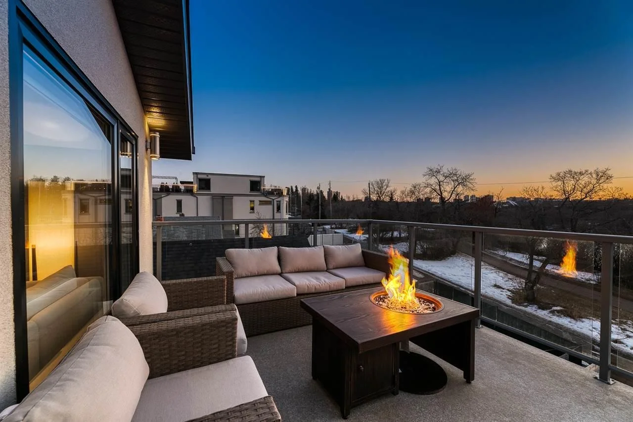 Outdoor balcony with cushioned wicker furniture and a fire pit during sunset, snow on the ground, trees, and neighboring buildings in the background.