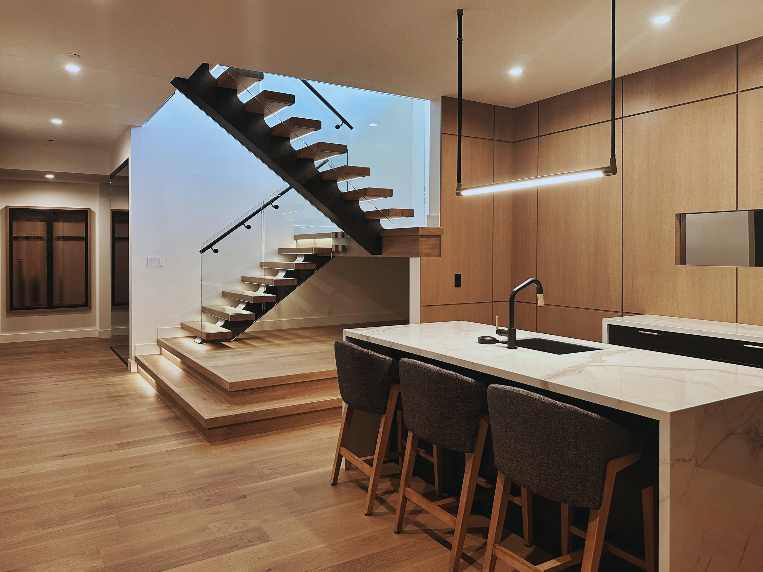 A modern kitchen featuring a marble countertop island with bar seating, wood cabinetry, and a glass-paneled staircase leading to an upper level. The space has warm wooden flooring and ambient lighting.