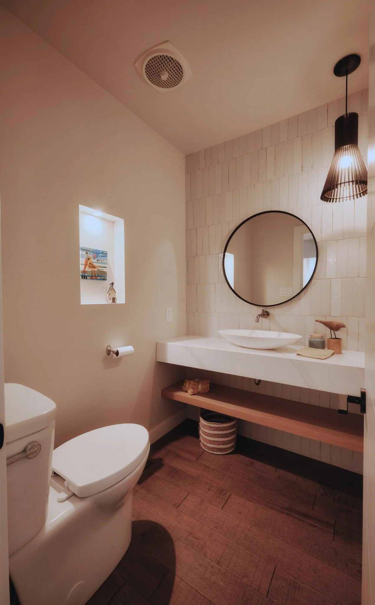 Modern bathroom with a white toilet, a floating marble sink with a vessel basin, a round mirror, a black hanging light, a small recessed shelf with decorative items, and wood flooring.