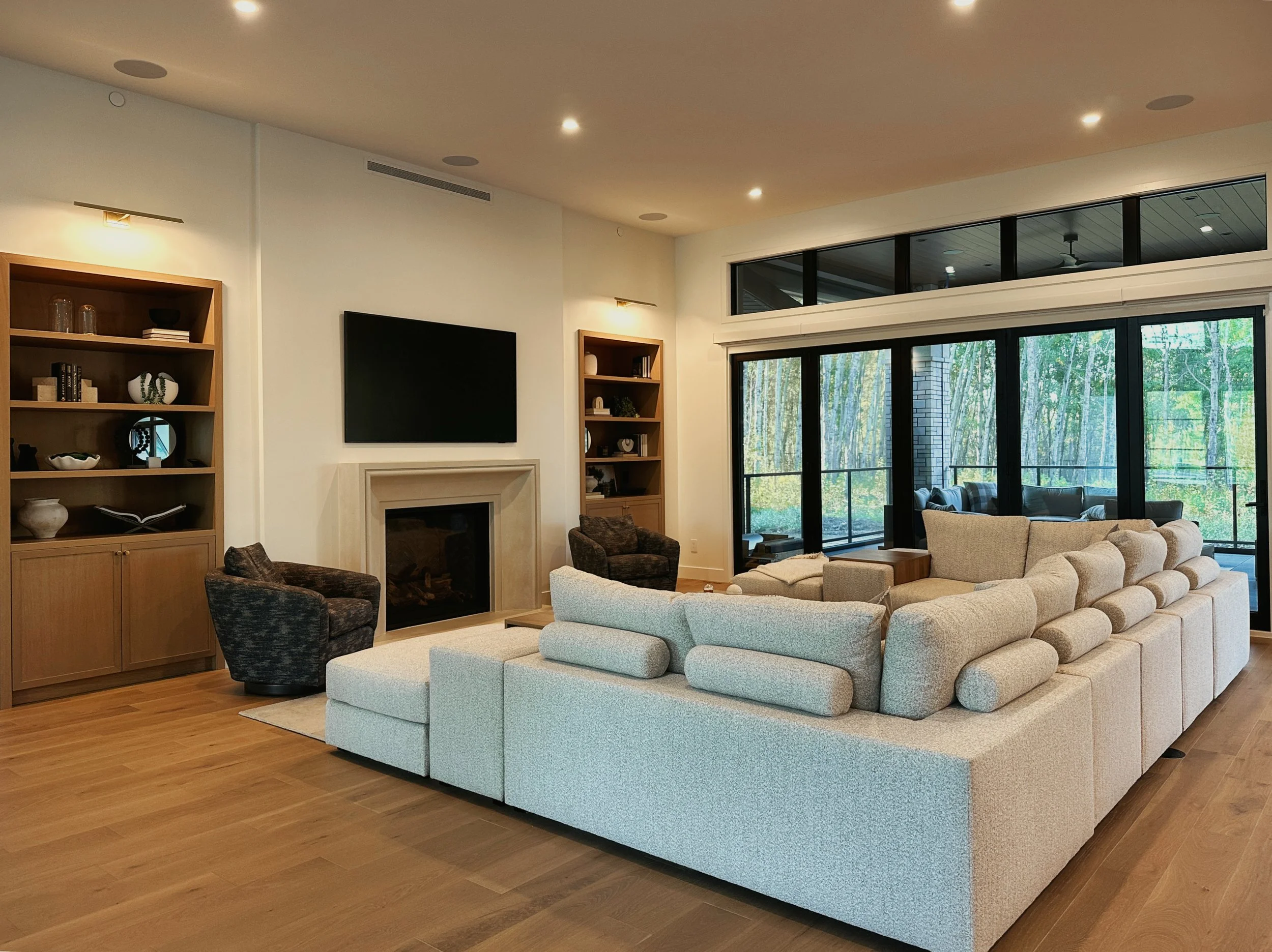 Modern luxury great room with large light blue modular sectional sofa, stone fireplace, wall-mounted TV, walnut bookshelves, massive black-framed sliding doors to forest view, high ceiling with recessed lights.