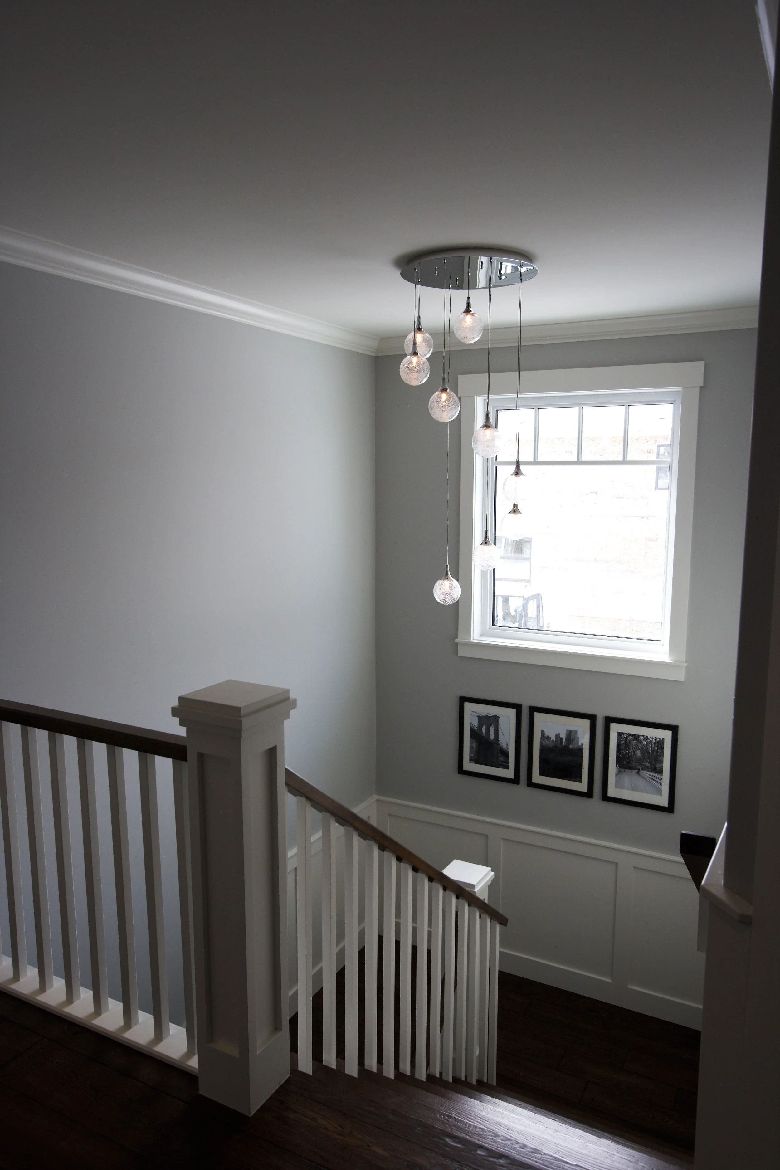 Interior view of a staircase landing with a modern chandelier hanging from the ceiling, a window, and three black-and-white framed photos on the wall.