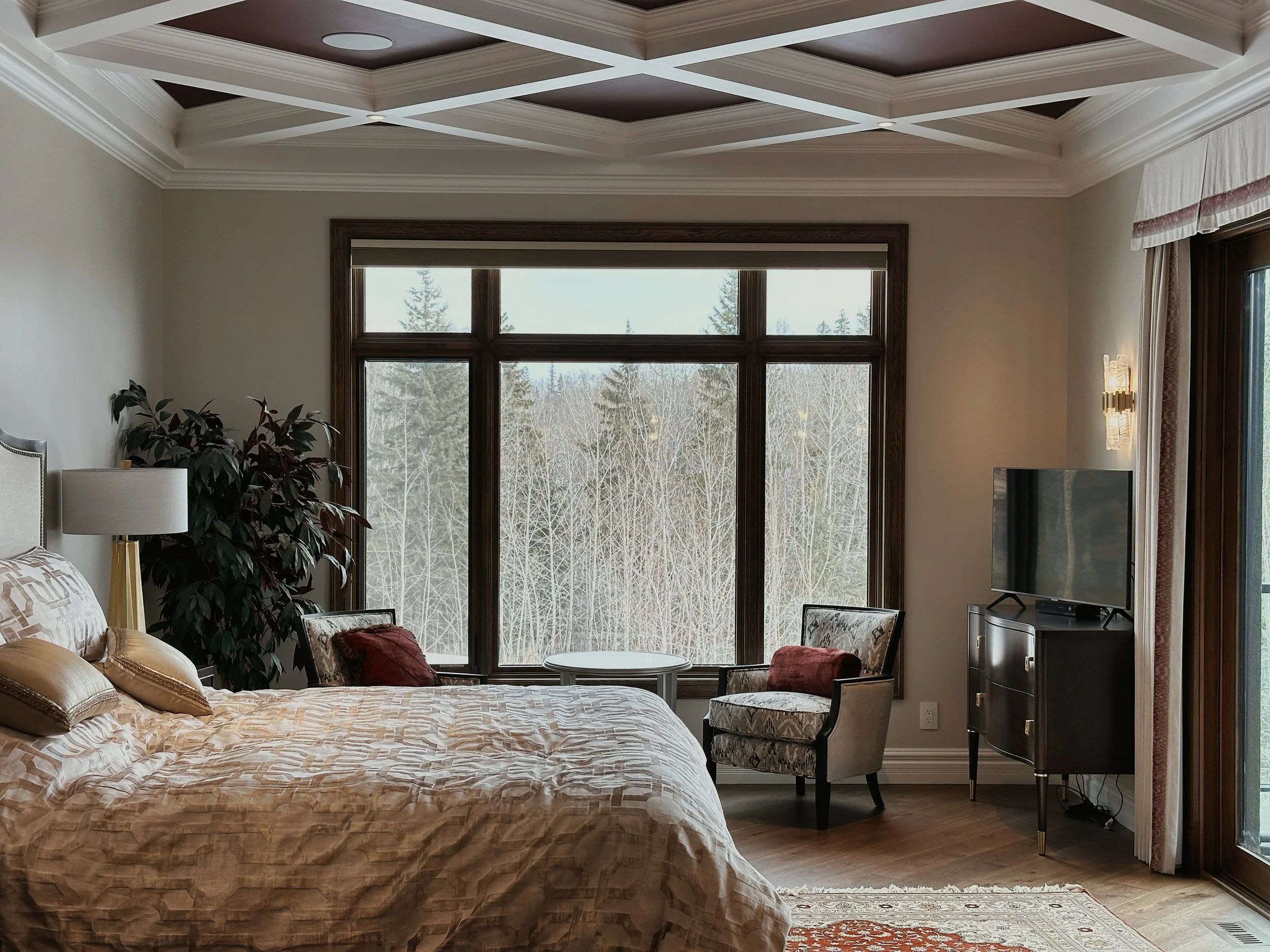A bedroom with a large window overlooking a wooded outdoor landscape, featuring a beds with beige patterned bedding, a bedside lamp, a potted plant, a chair with pillows, a small table, a dresser with a television, and a coffered ceiling.