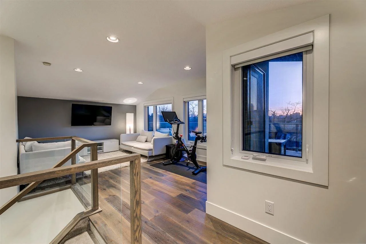 Living room with a wall-mounted TV, white couches, exercise bike near large windows, and a view of the sunset outside.