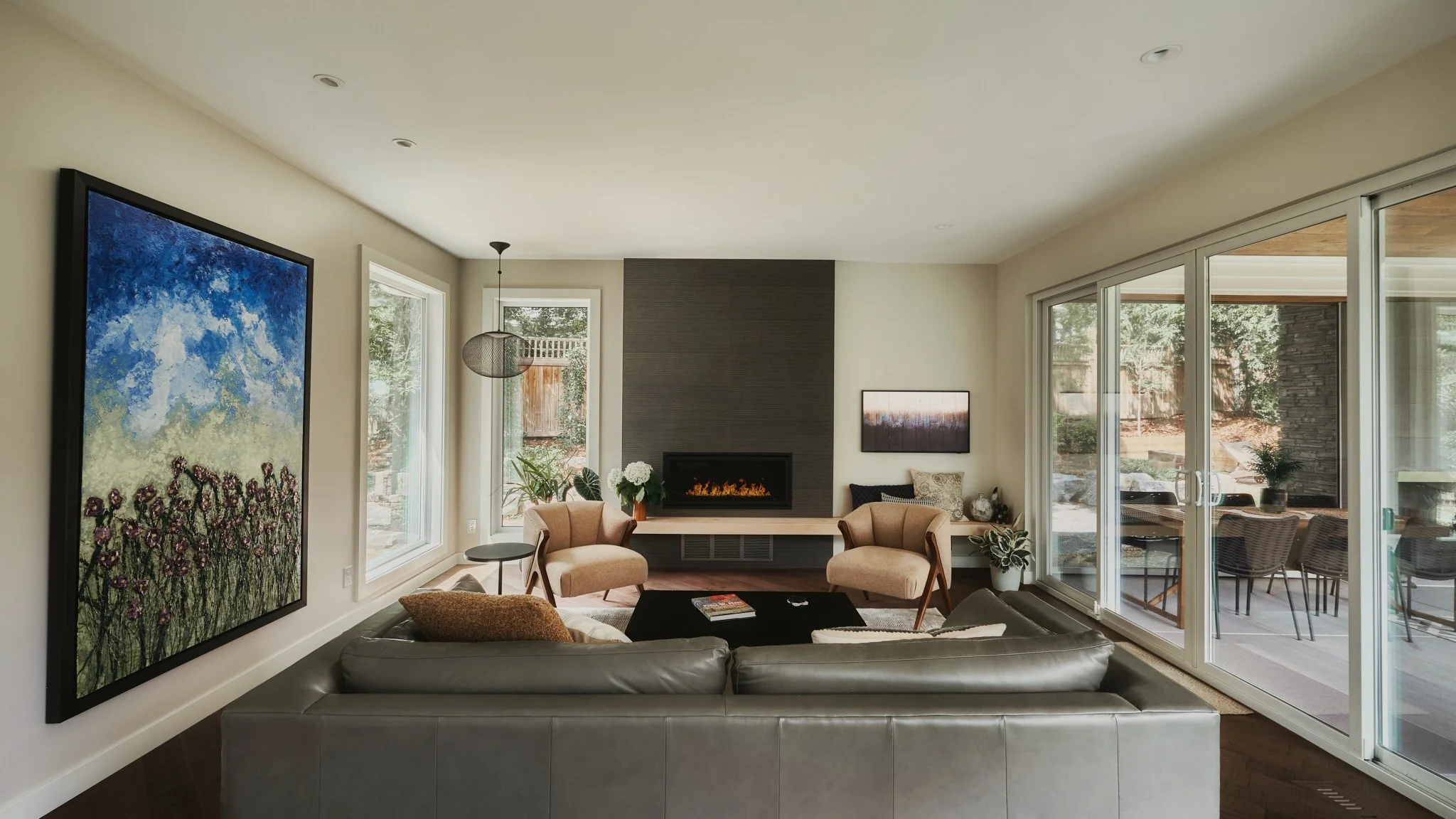 Modern living room with large sliding glass doors opening to a patio, two beige armchairs near a fireplace, a gray sofa, a black coffee table, and wall-mounted TV.