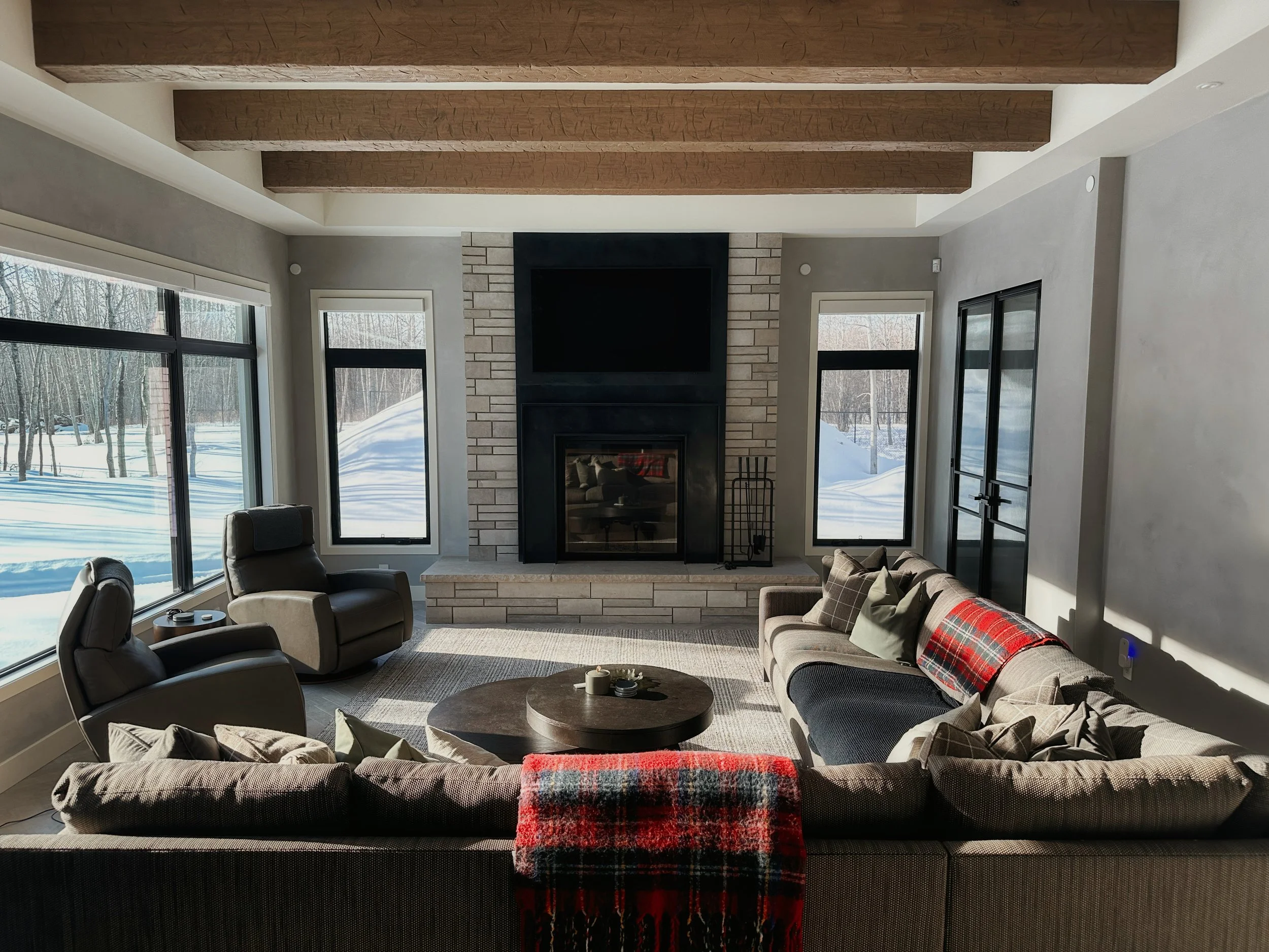 High-end rustic-modern family room with spacious sectional sofa in earthy tones and plaid blanket, gray recliner chairs, round dark wood coffee table, prominent stone-clad fireplace with TV above, vaulted ceiling with exposed beams.