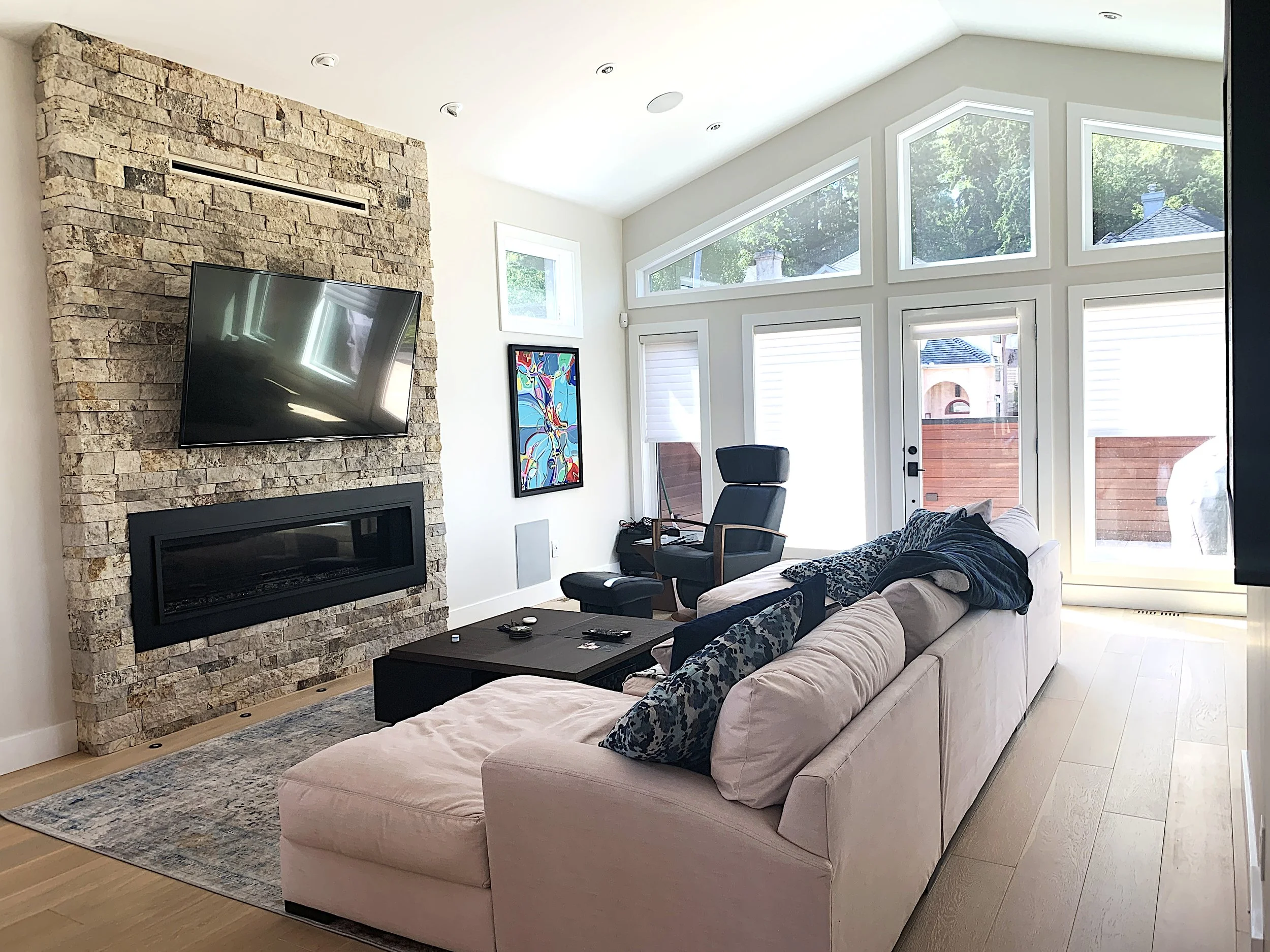 Modern living room with vaulted ceiling, stone accent wall, fireplace, and large windows—features sectional sofa, abstract art, and natural light, ideal for custom home builder luxury interiors.