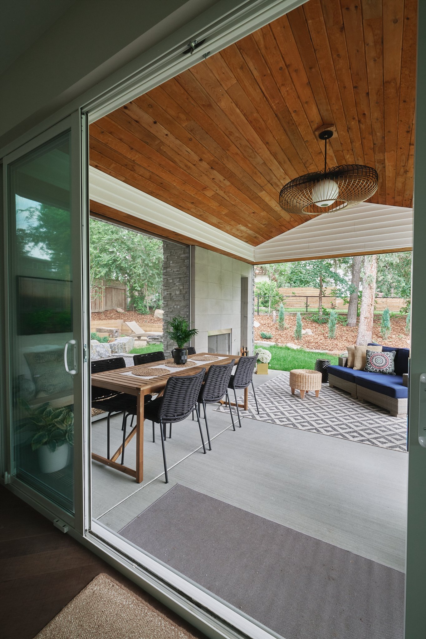 Covered patio with wood ceiling, dining table, woven chairs, and cozy seating area overlooking backyard.
