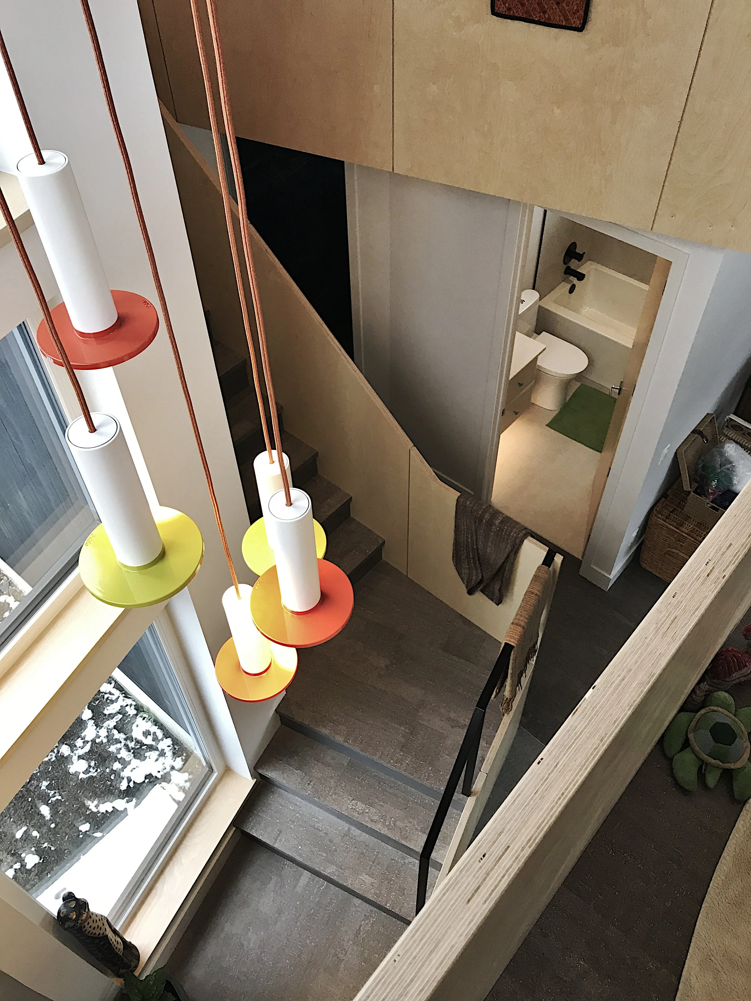 Modern home interior with staircase, wood-paneled walls, colorful hanging light fixture, and bathroom view—features natural light, snowy window scene, and minimalist design with cozy decorative accents.