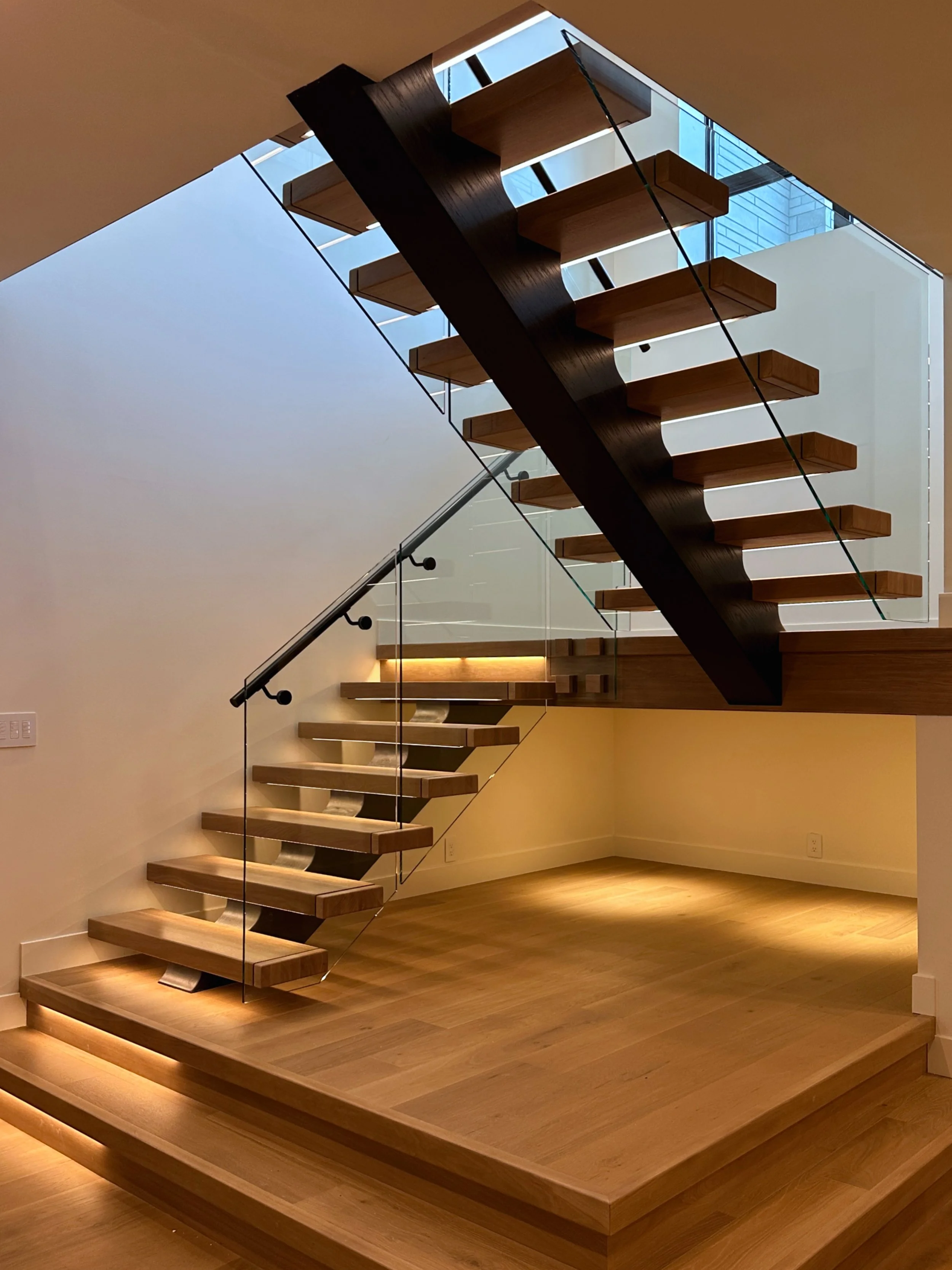 High-end modern open-riser staircase with cantilevered dark walnut steps, glass side support, black minimalist handrail, glowing LED illumination on each tread, matching light hardwood flooring.