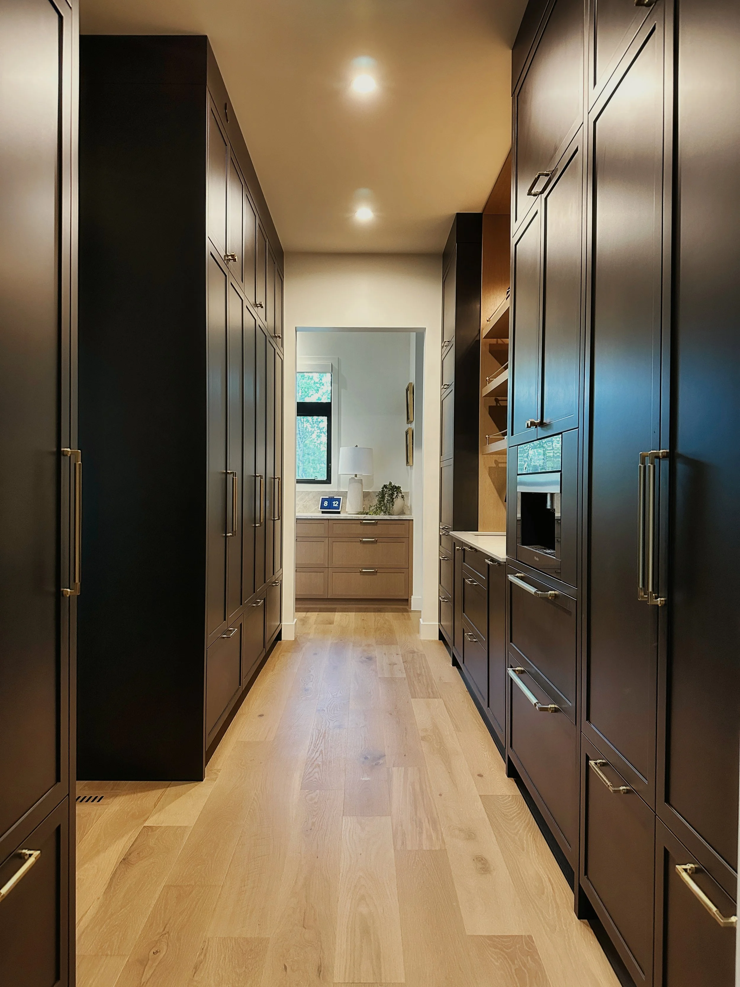 High-end contemporary butler’s pantry featuring tall charcoal cabinets, brass pulls, quartz countertops, integrated oven and microwave, floating shelves, warm wood flooring, pot lights, and open doorway to kitchen.