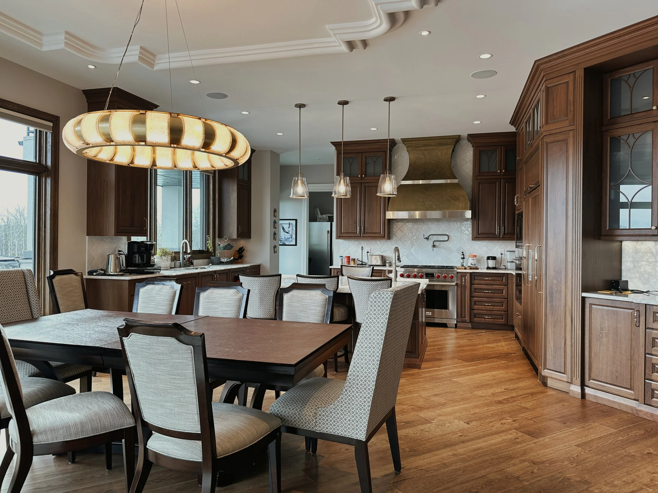 Spacious kitchen with custom wooden cabinets, dining table with upholstered chairs, pendant lighting, plaster ceiling detail and large windows.