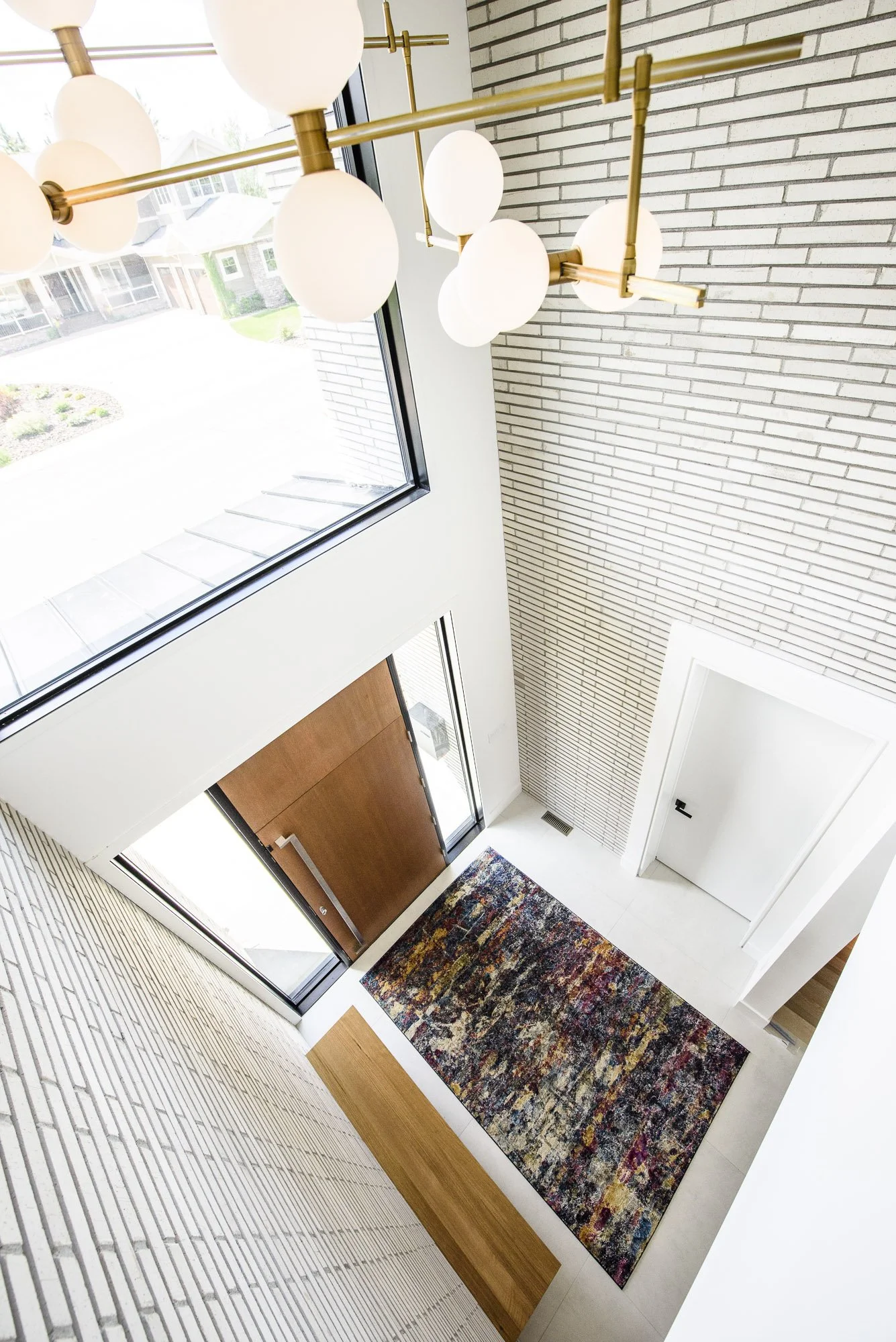 Open-concept double-height foyer design with statement brass and glass chandelier, whitewashed brick walls, transom and sidelight windows flooding space with daylight, rich wood entry door, and bold abstract multicolored rug.