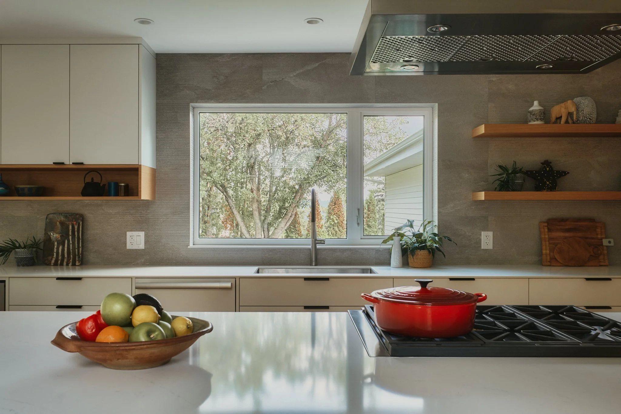 Modern kitchen with white countertops, a bowl of assorted fruits, a red pot on the stove, and a large window looking outdoors with trees.