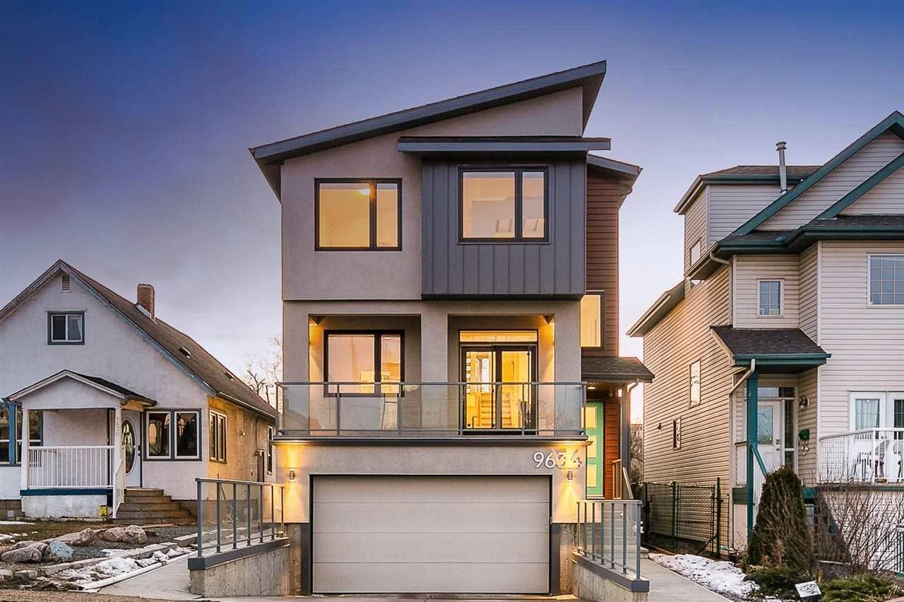 Modern three-story house with large windows and a garage, situated between two older houses, during twilight with a clear sky.