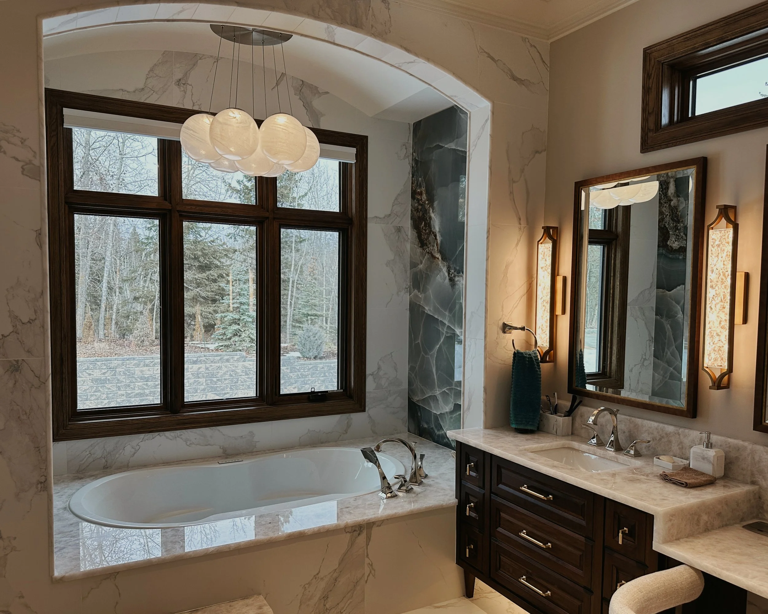 Luxury bathroom with a bathtub under a large window, marble walls, a double sink vanity with mirrors, and modern lighting fixtures.