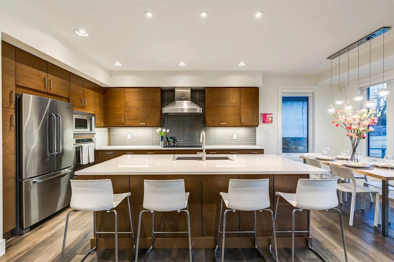 Modern kitchen with wooden cabinets, stainless steel refrigerator, built-in oven, gray backsplash, white countertop island with sink, four white bar stools, adjacent dining area with a wood table set for dinner, floral centerpiece, large sliding glass door, and pendant lighting.